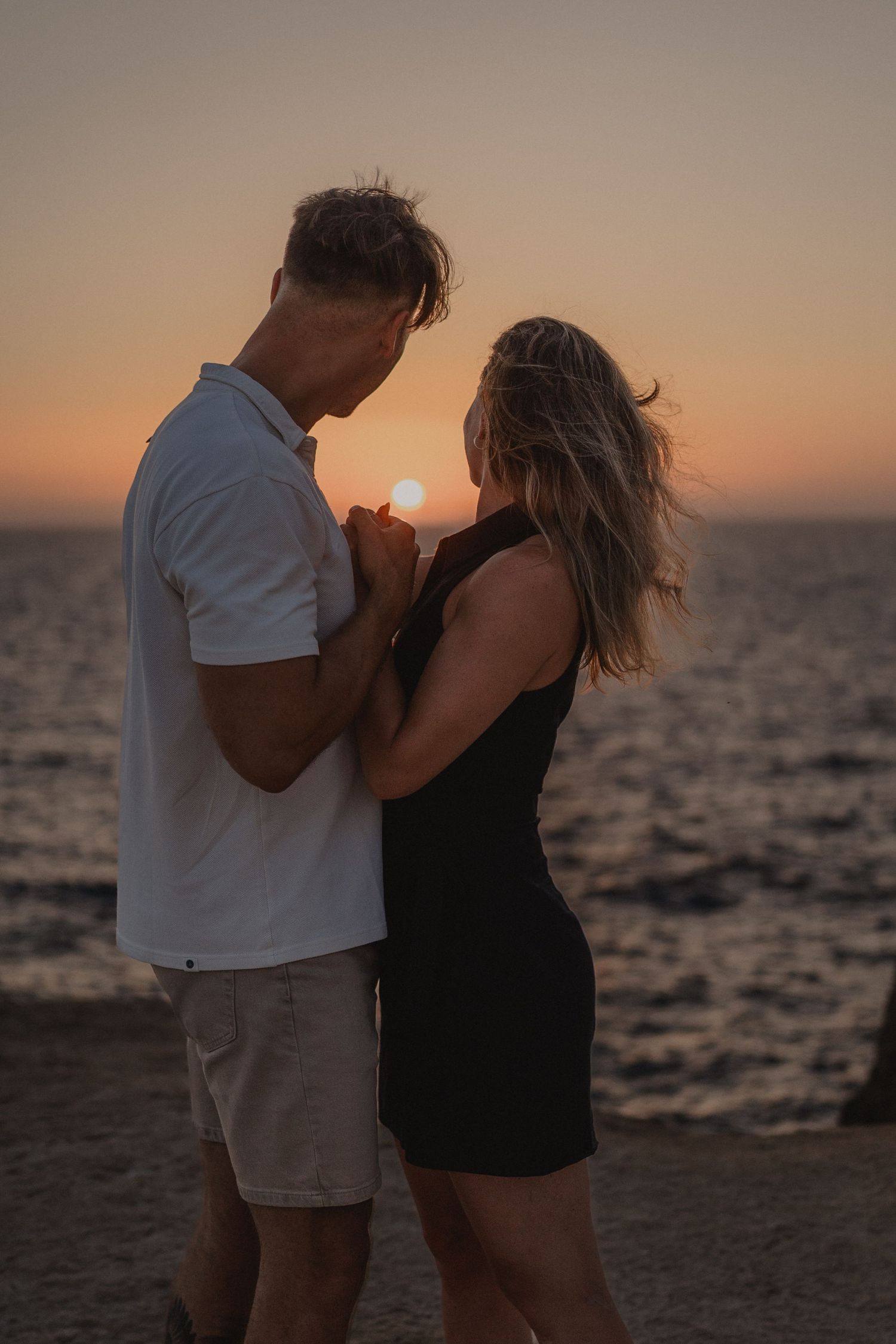 Romantic couple holding hands at sunset by the ocean, woman in black dress and man in white polo shirt.