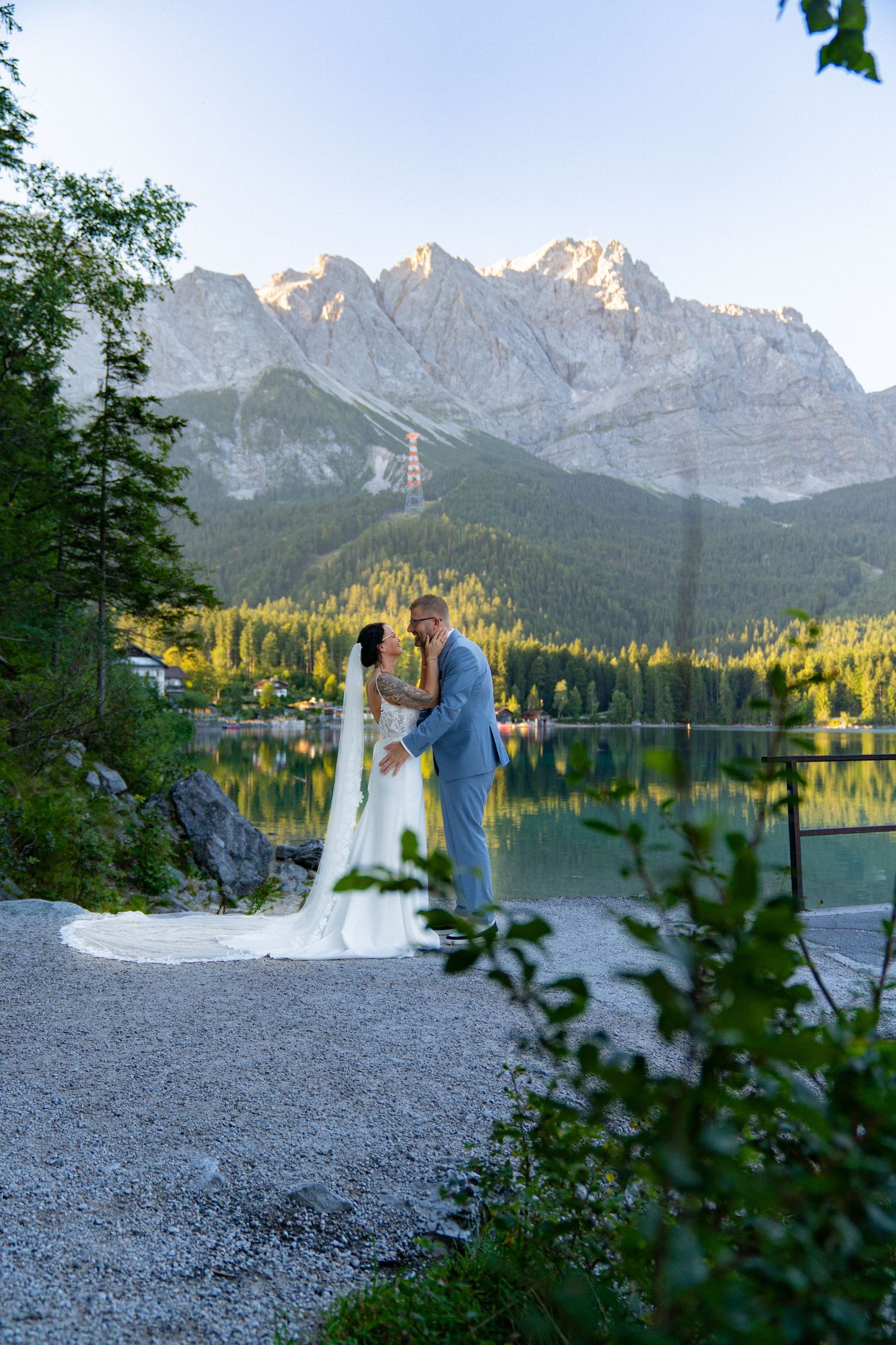 A couple shares a romantic moment by a serene mountain lake at sunset with majestic peaks in the background.