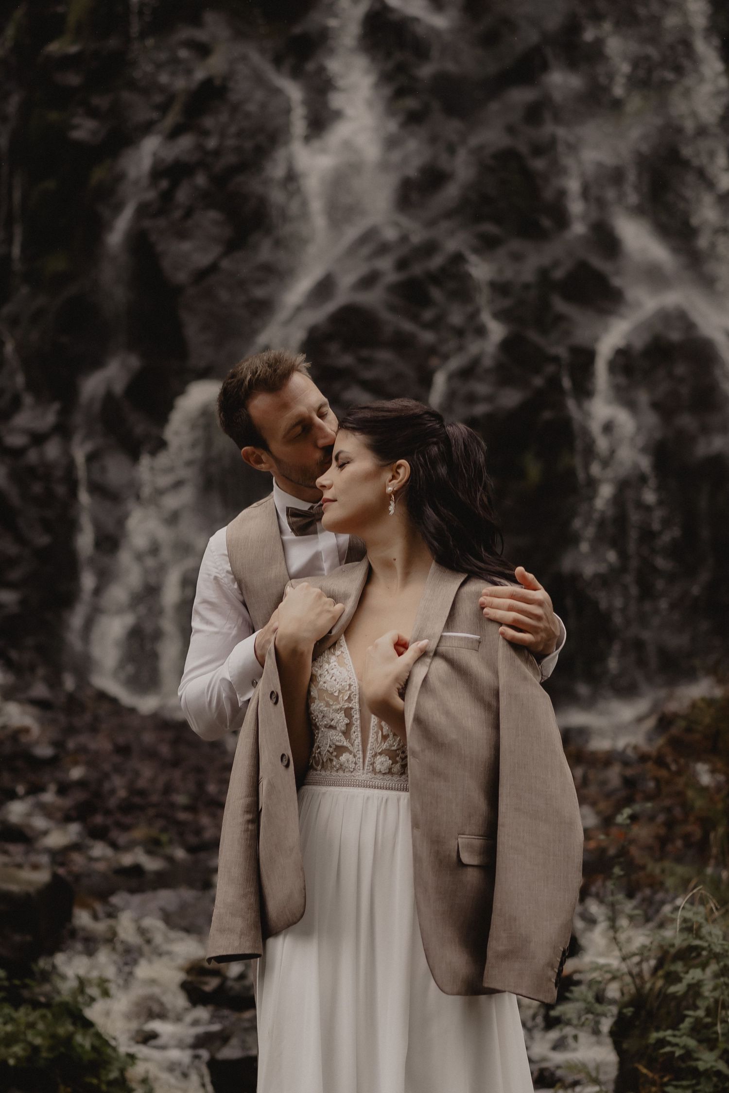 A romantic couple embraces near a dramatic waterfall backdrop in moody lighting.