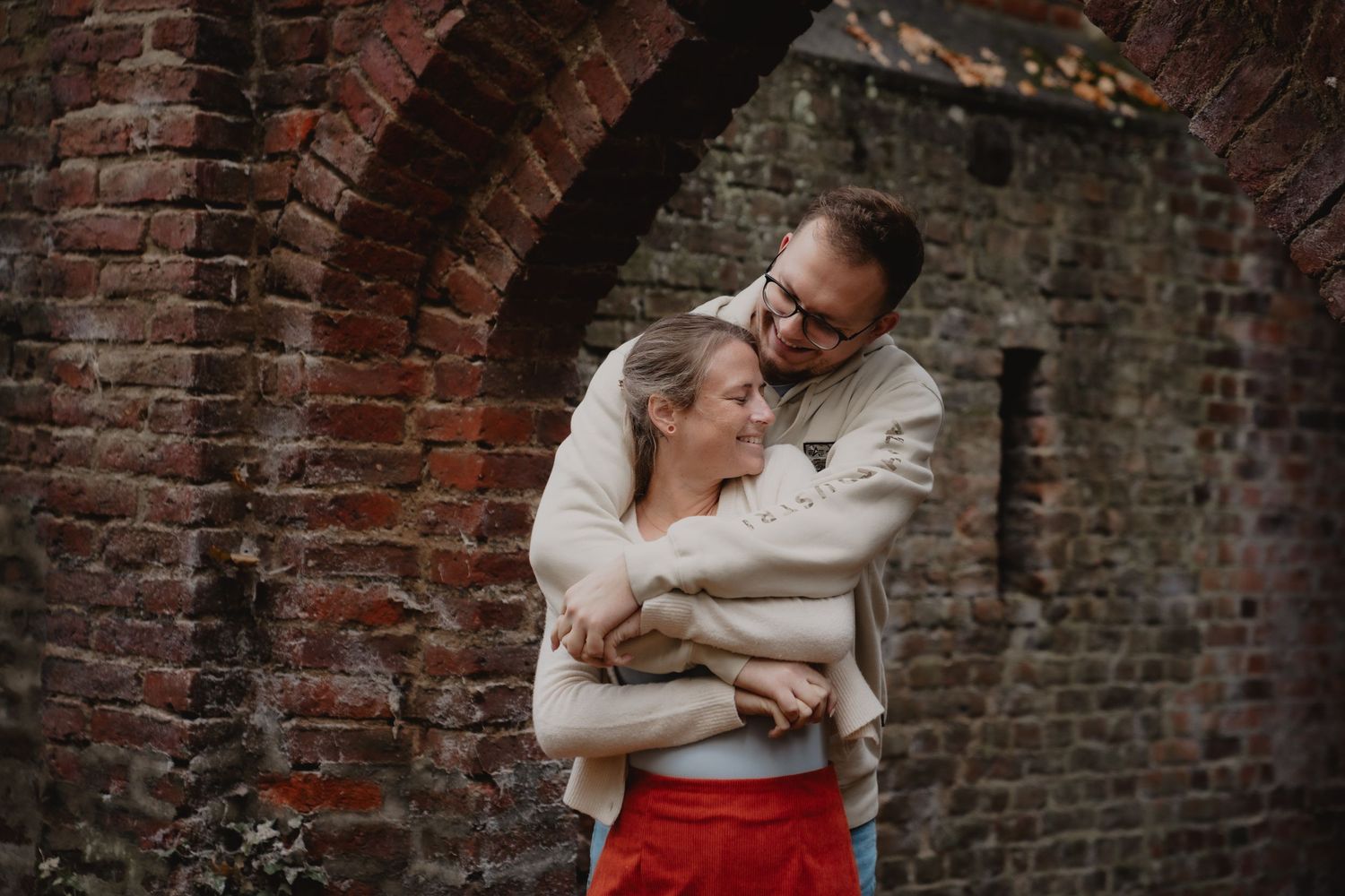 A couple embraces against a rustic brick wall background during an outdoor photoshoot.