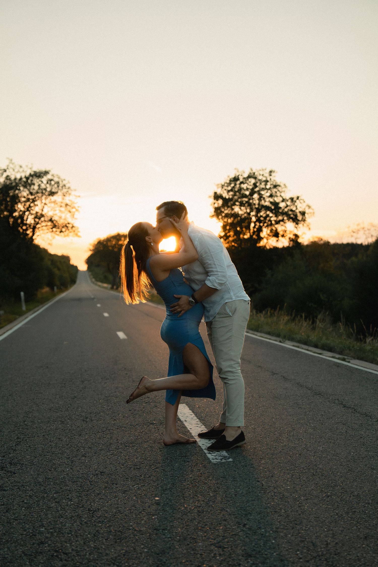 Couple sharing a romantic kiss on an empty road at golden sunset, surrounded by trees in warm evening light.