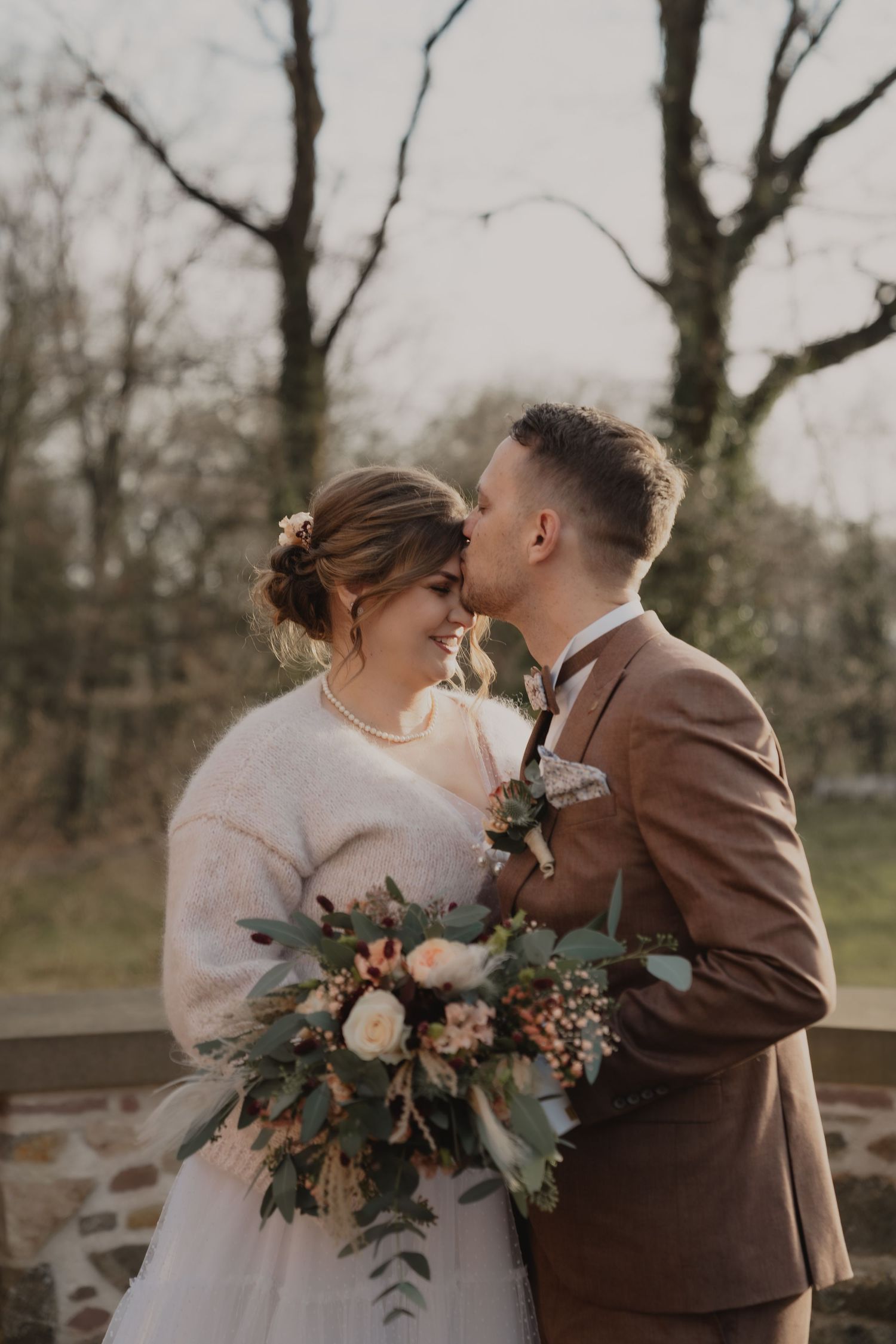 A romantic wedding couple embraces while holding a floral bouquet outdoors.