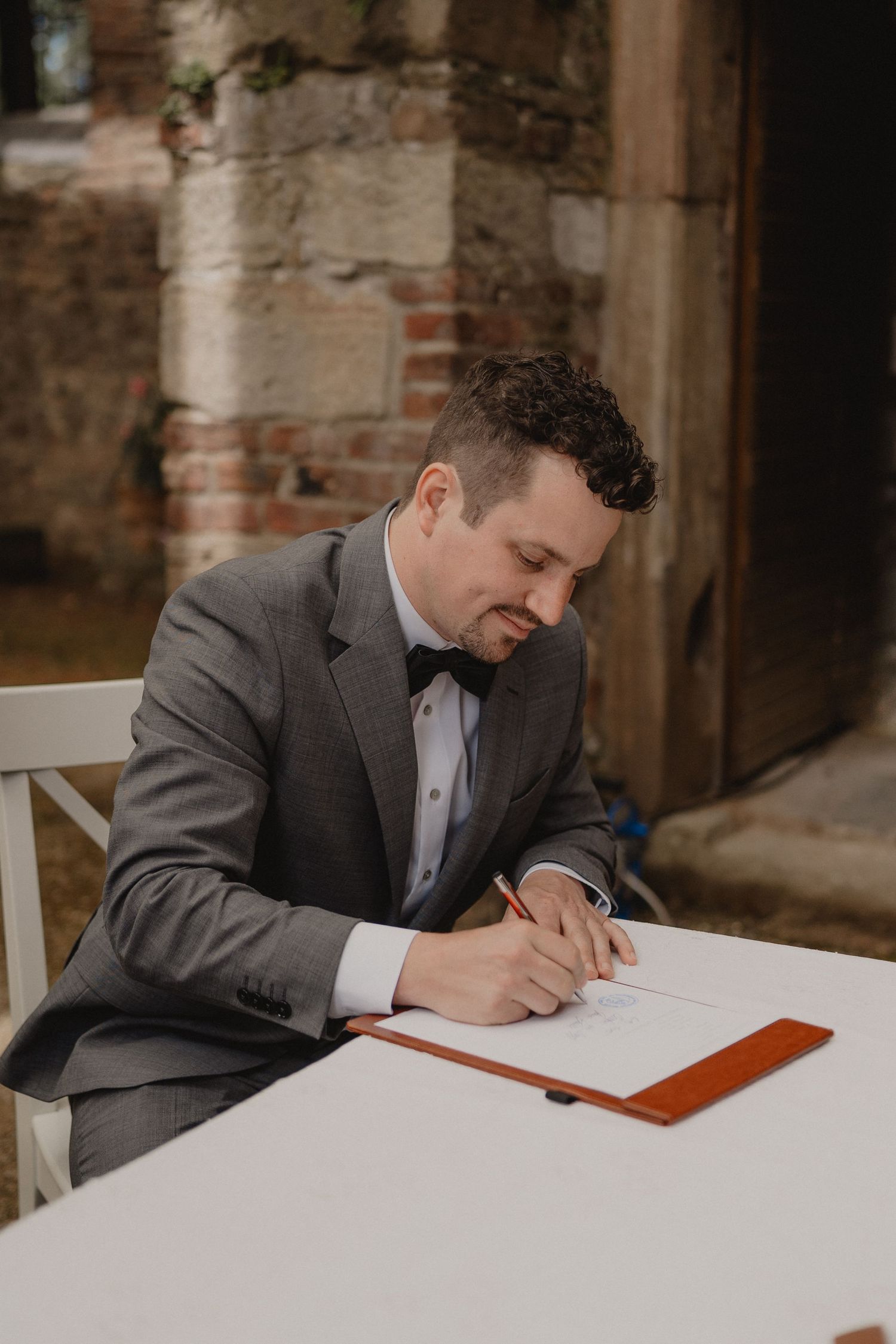 Person in formal attire signs marriage documents at a white table against a rustic brick wall backdrop.