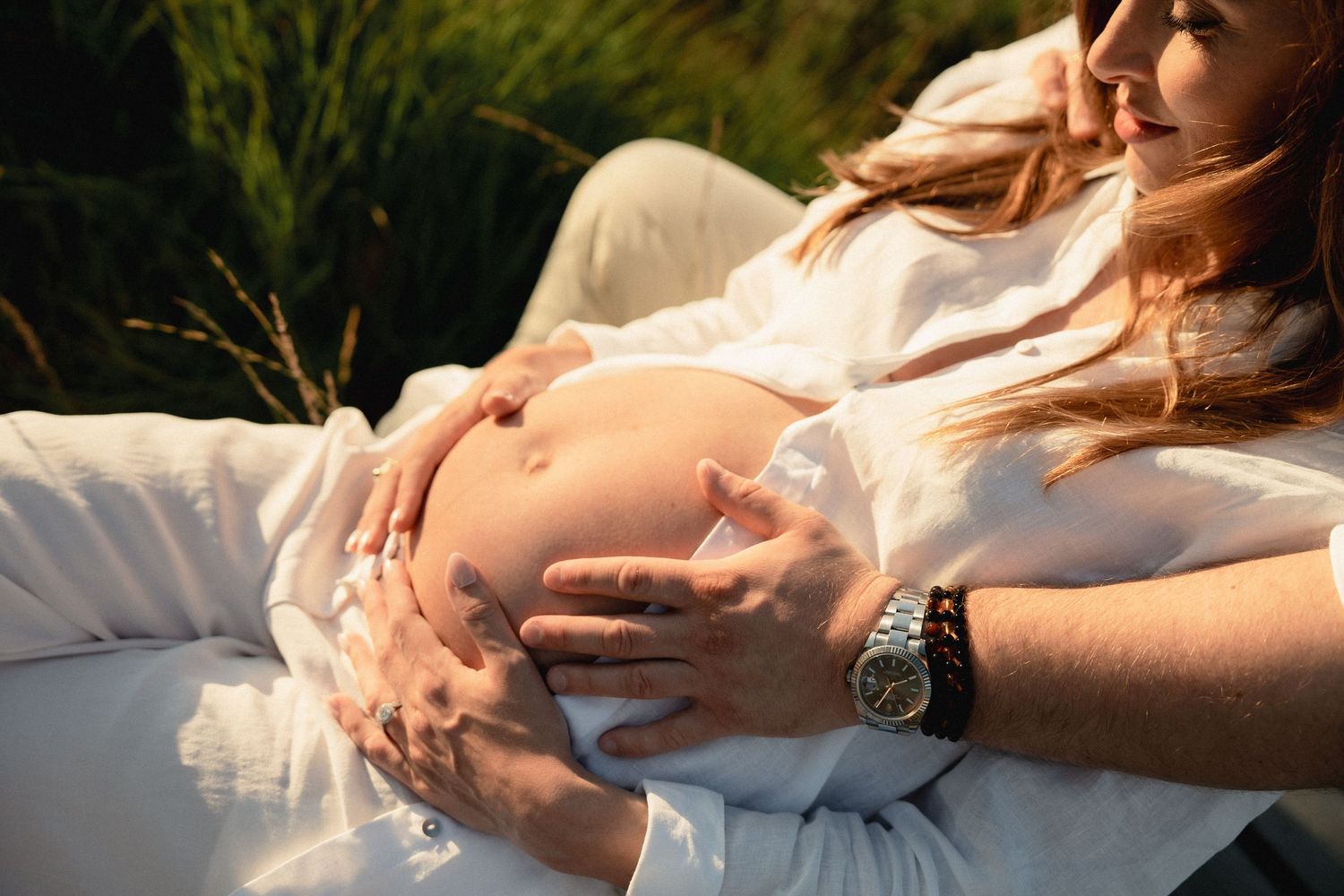 Couple in white clothing outdoors, pregnant woman cradling baby bump with partner's hands resting gently on belly.