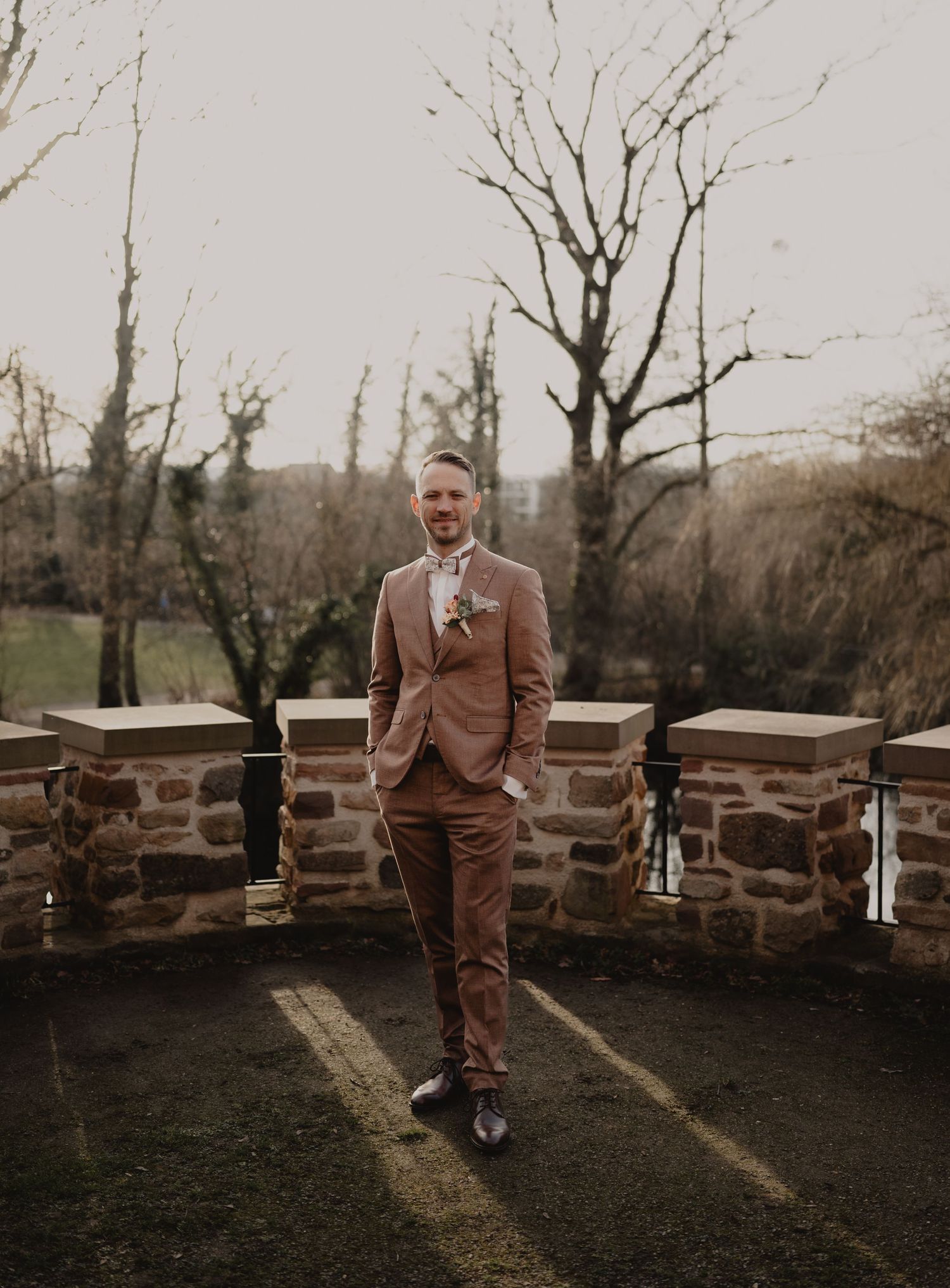A person in a brown suit stands in front of a rustic stone wall on a moody autumn day.