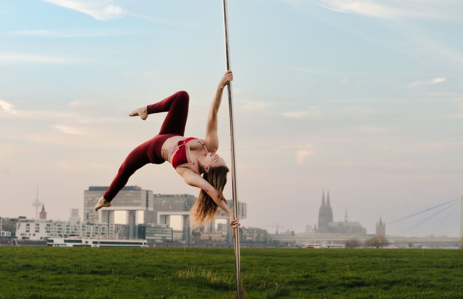Woman performing pole dance outdoors on green field with Cologne Cathedral skyline in background.