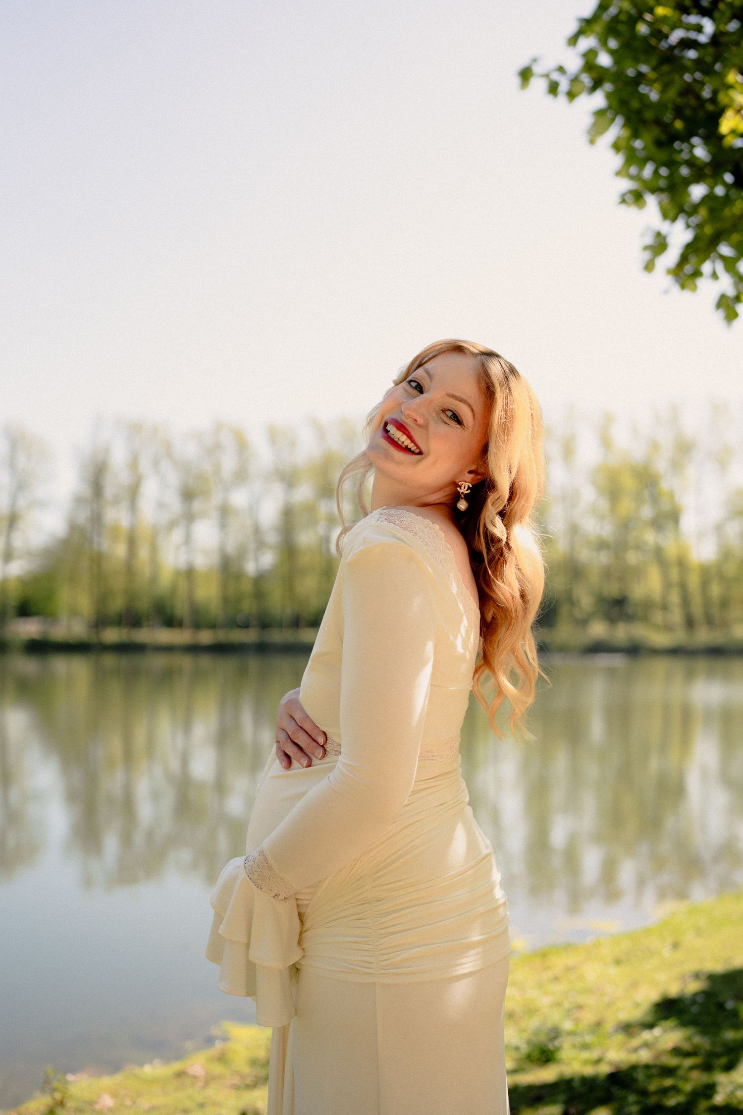 Radiant pregnant woman in cream dress smiling by a reflective lake at golden hour.