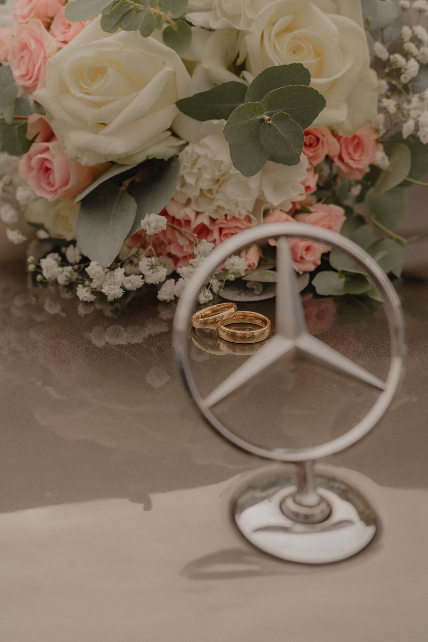 Wedding rings displayed on a Mercedes-Benz emblem with roses and eucalyptus in the background.