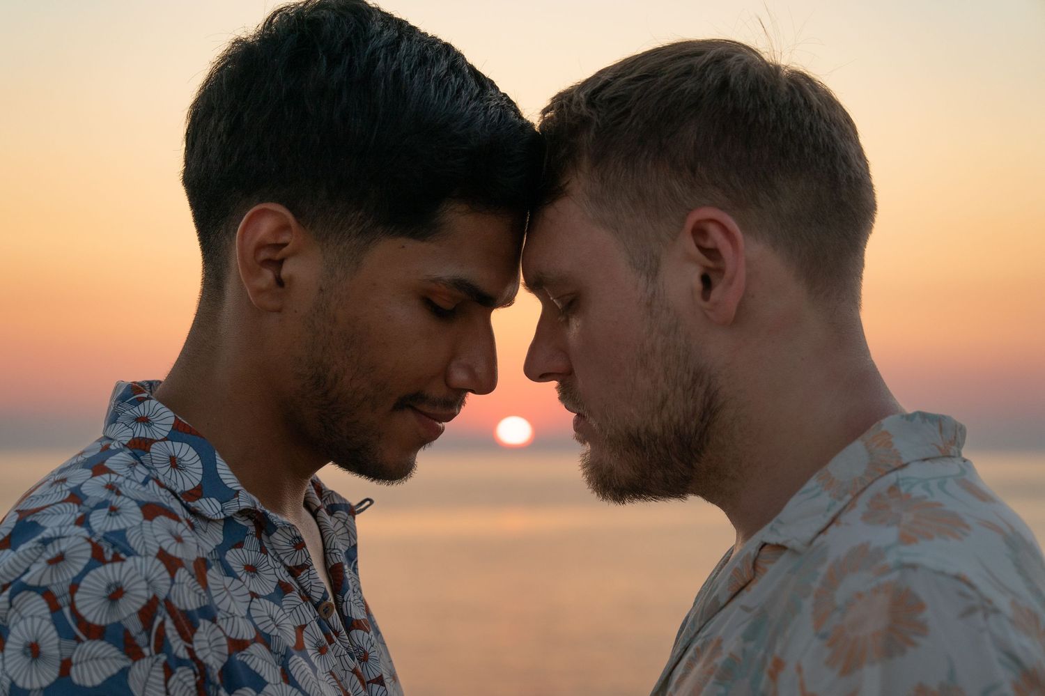 Two young men touch foreheads romantically at sunset by the ocean, wearing floral shirts in warm golden light.