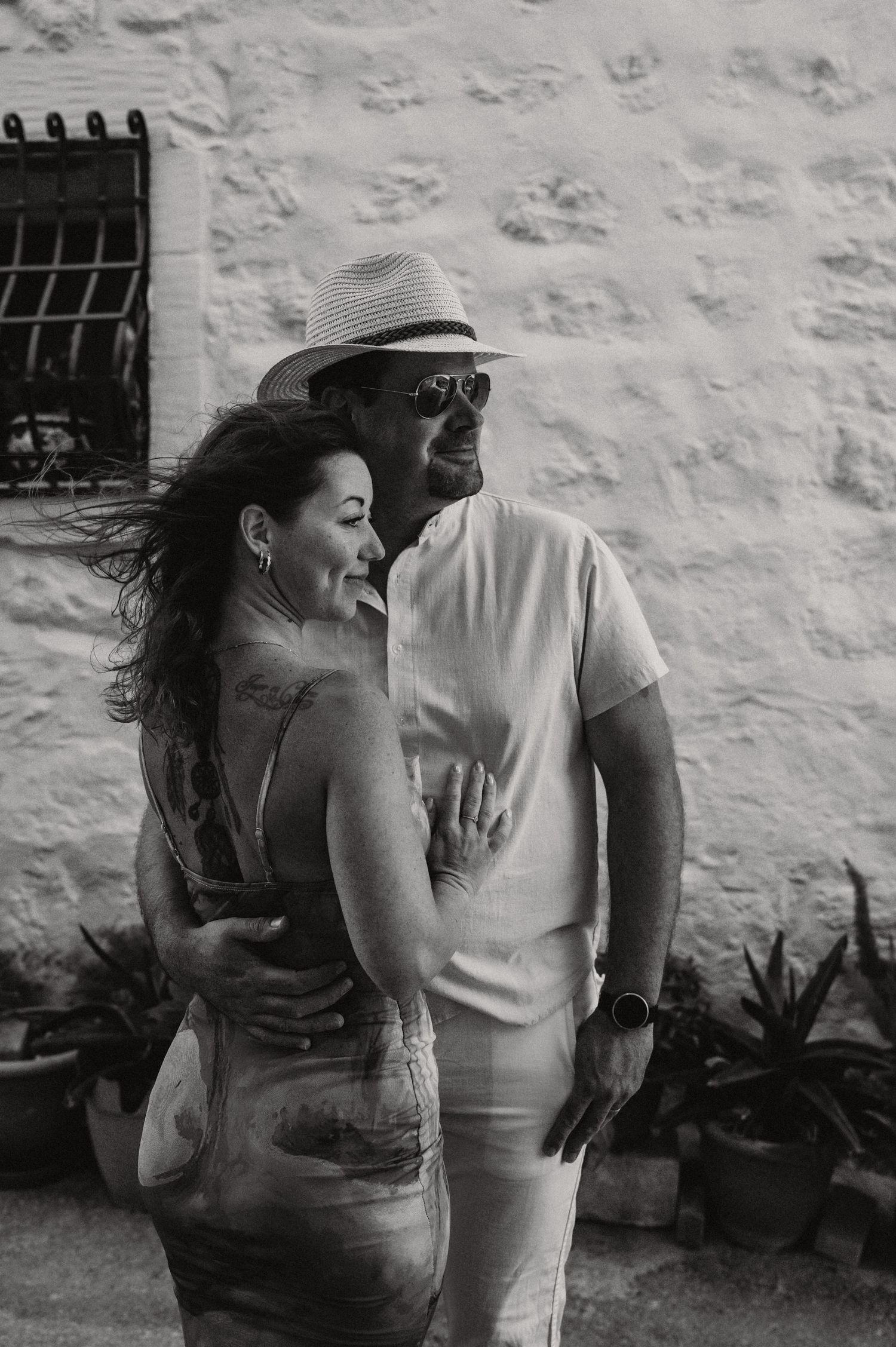 Black and white photo of a couple embracing and smiling outdoors near a white wall with potted plants.