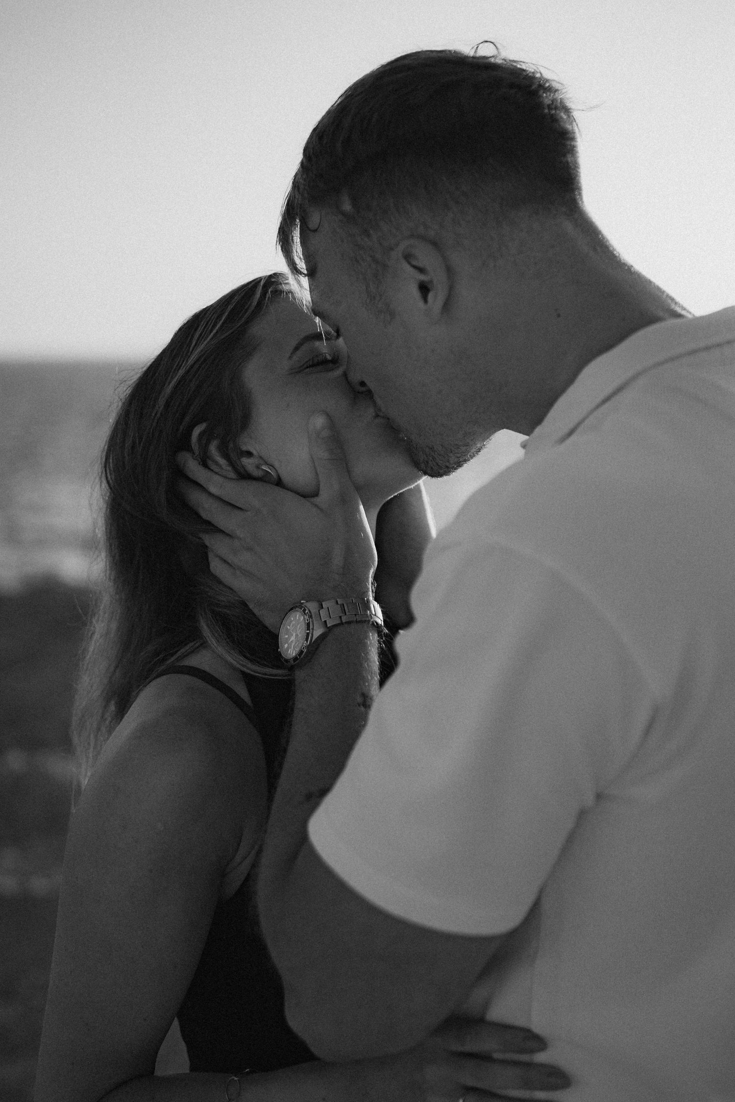 Black and white photo of a couple sharing a romantic kiss at sunset near the ocean, man tenderly holding womans face.