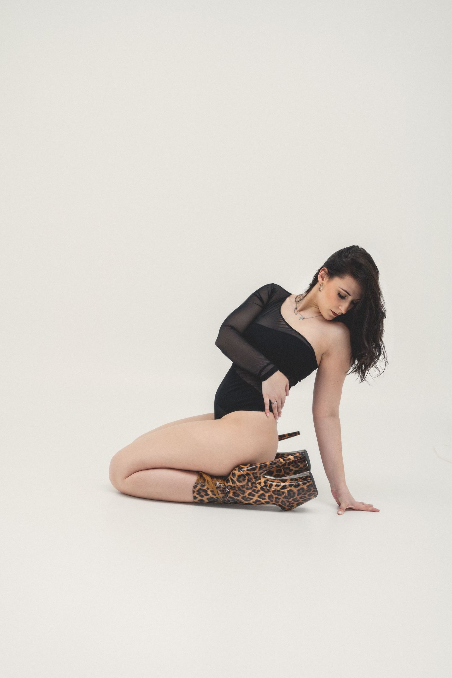 Woman kneeling in black mesh bodysuit holding leopard print heels on white background.