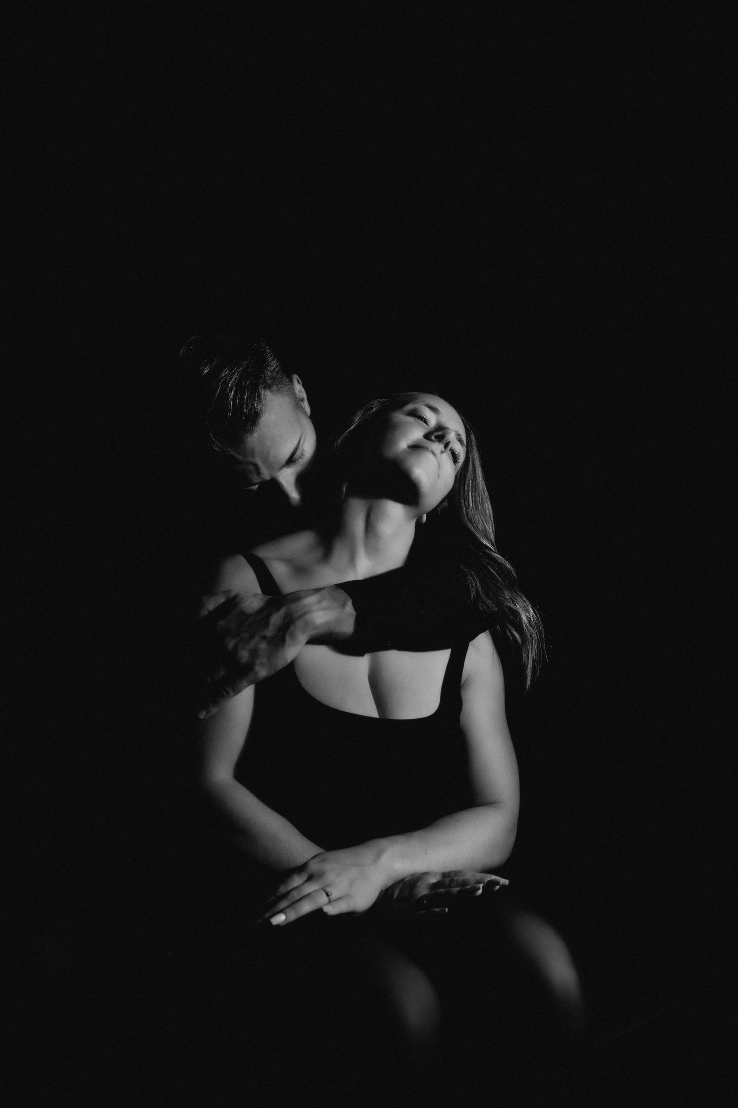 Black and white romantic couple portrait with dramatic lighting against dark background.