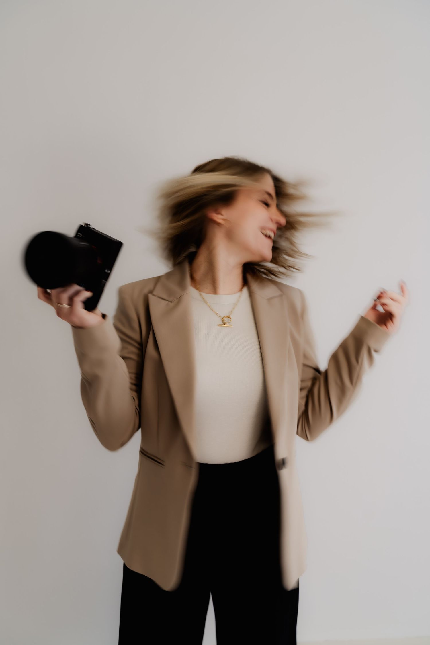 A series of dynamic fashion photos showing a person in a tan blazer and cream top with flowing hair motion against white wall.