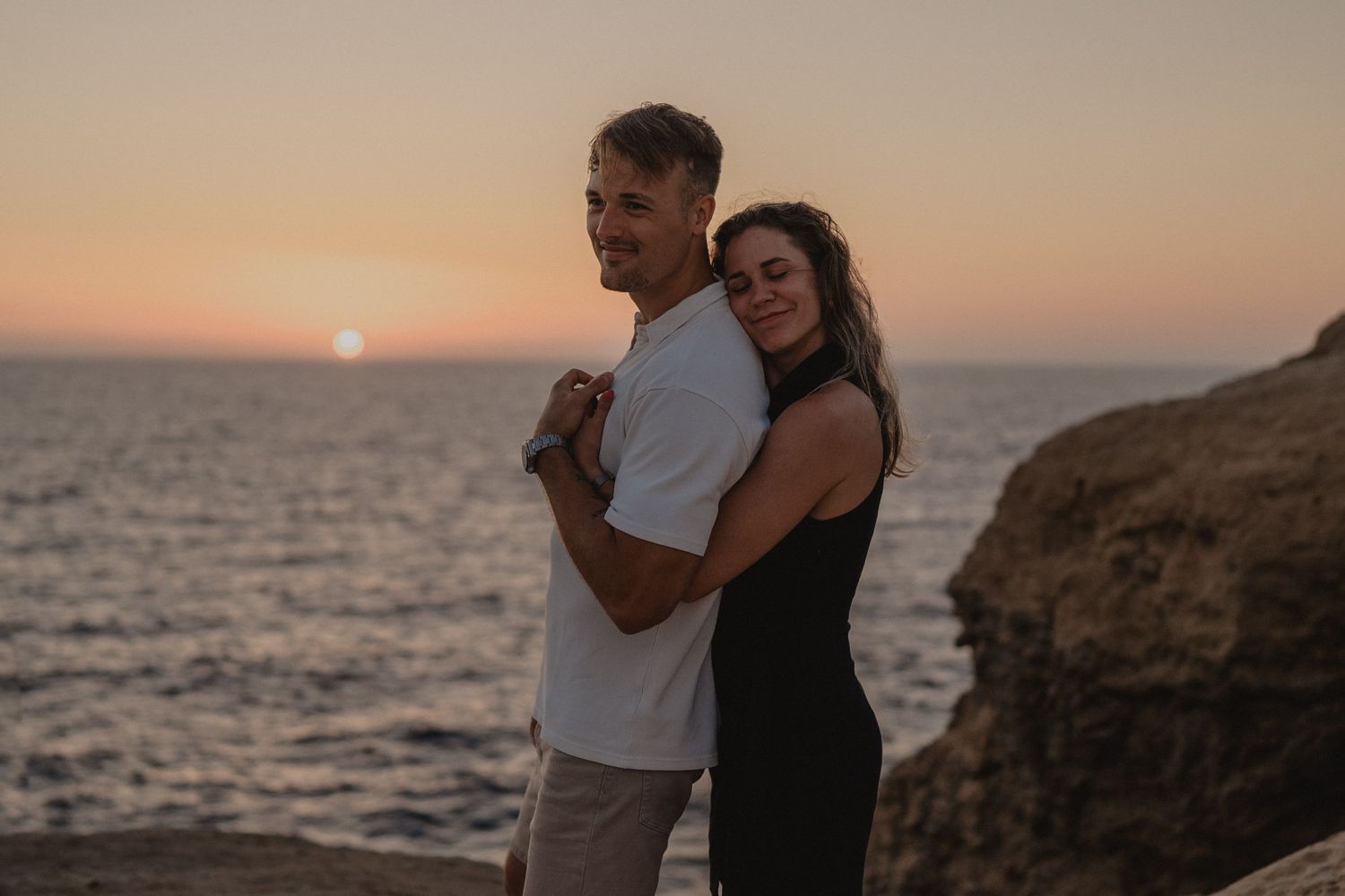 Couple embracing at sunset by the ocean, woman in black dress hugging man in white shirt near rocky cliffs.