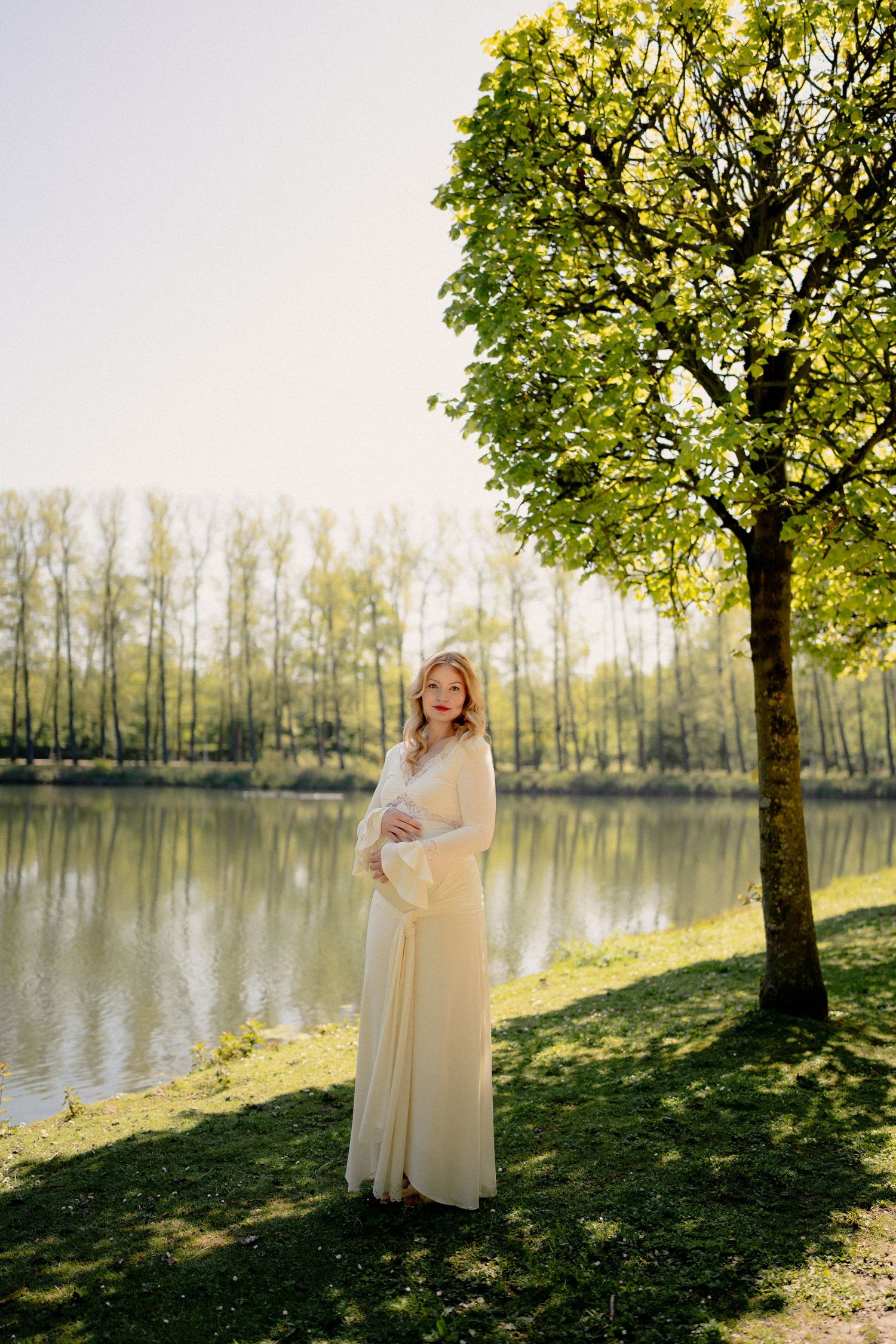 Pregnant woman in cream dress standing by a serene lake under a lush green tree in spring.