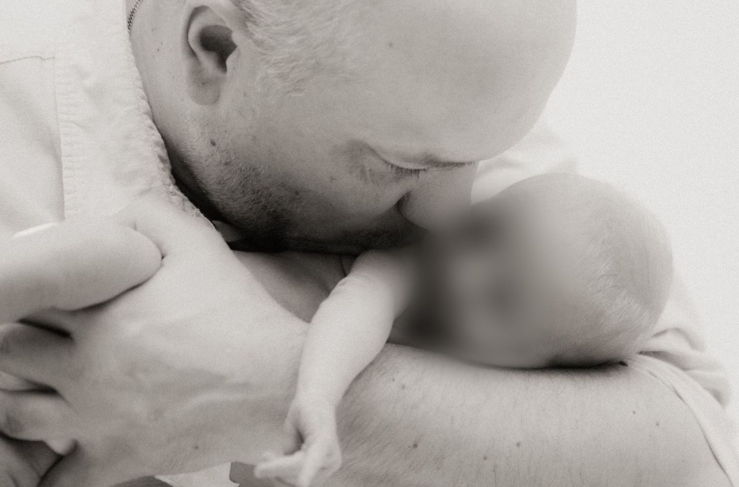 Black and white photo of a bald father tenderly kissing his newborn baby while cradling the infant in his arms.