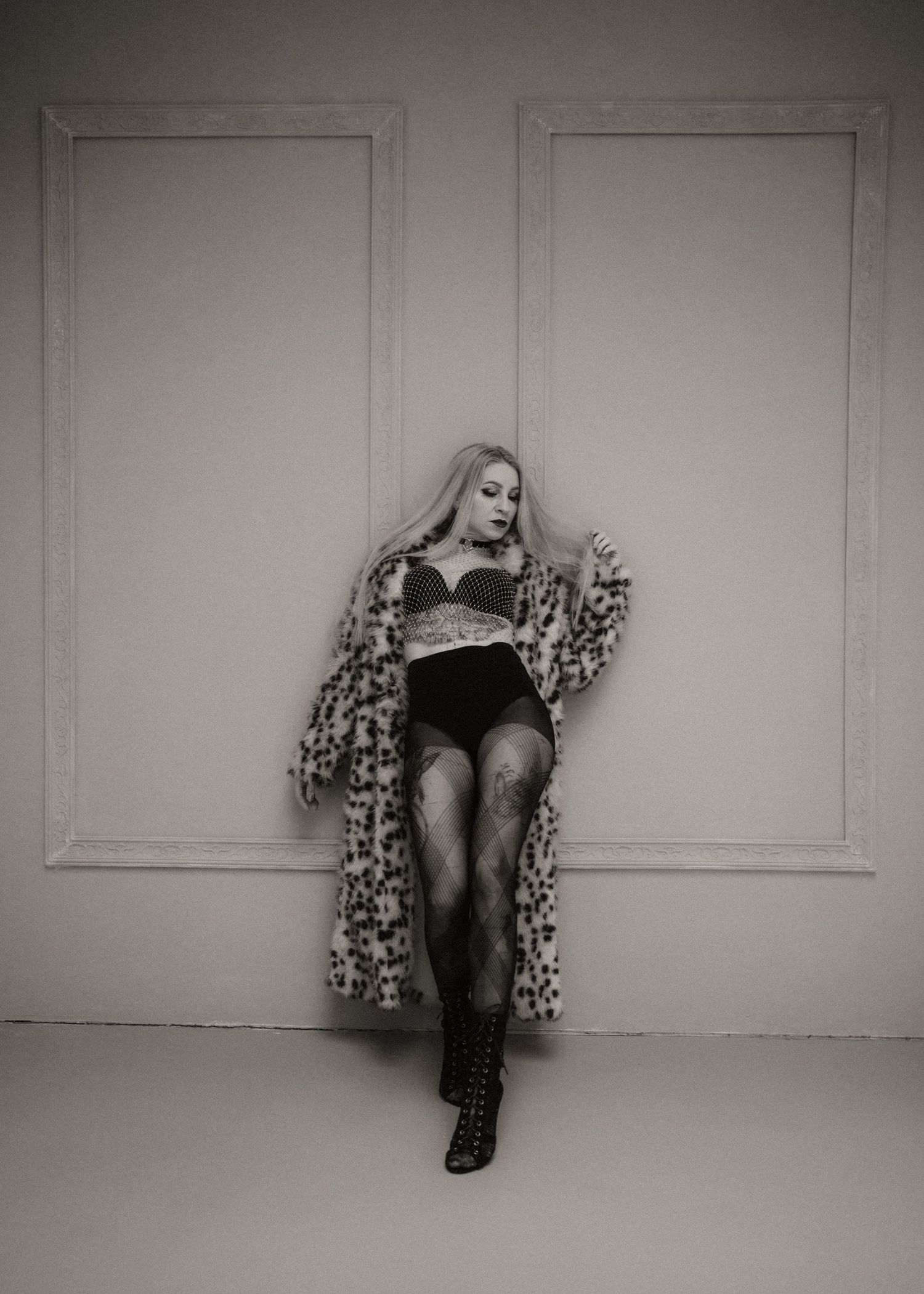 Black and white fashion photo showing a figure in lingerie and leopard print coat posing against a minimalist wall.