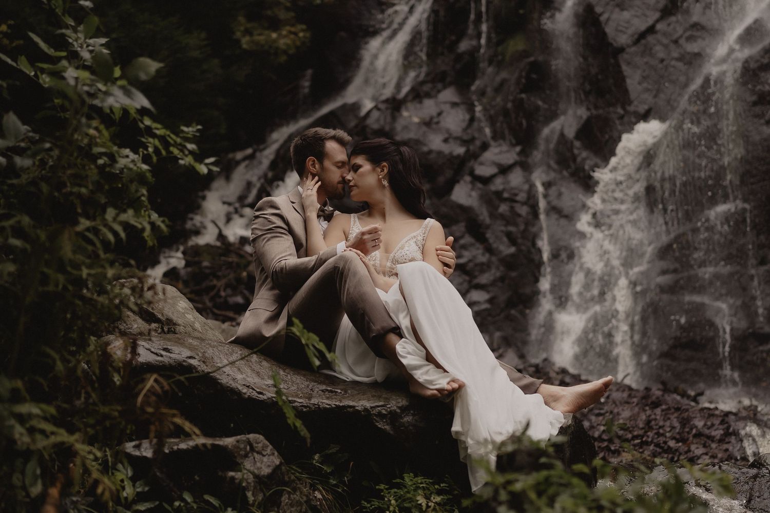 Couple in wedding attire embraces near a cascading waterfall in a lush forest setting.