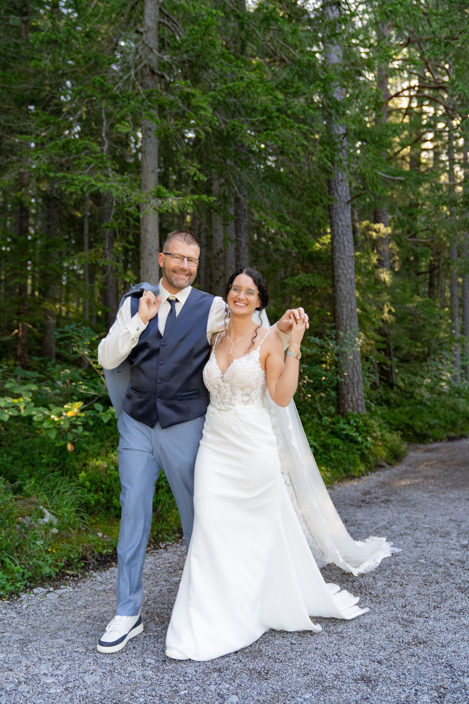 A couple in wedding attire playfully poses on a gravel path surrounded by lush green forest.