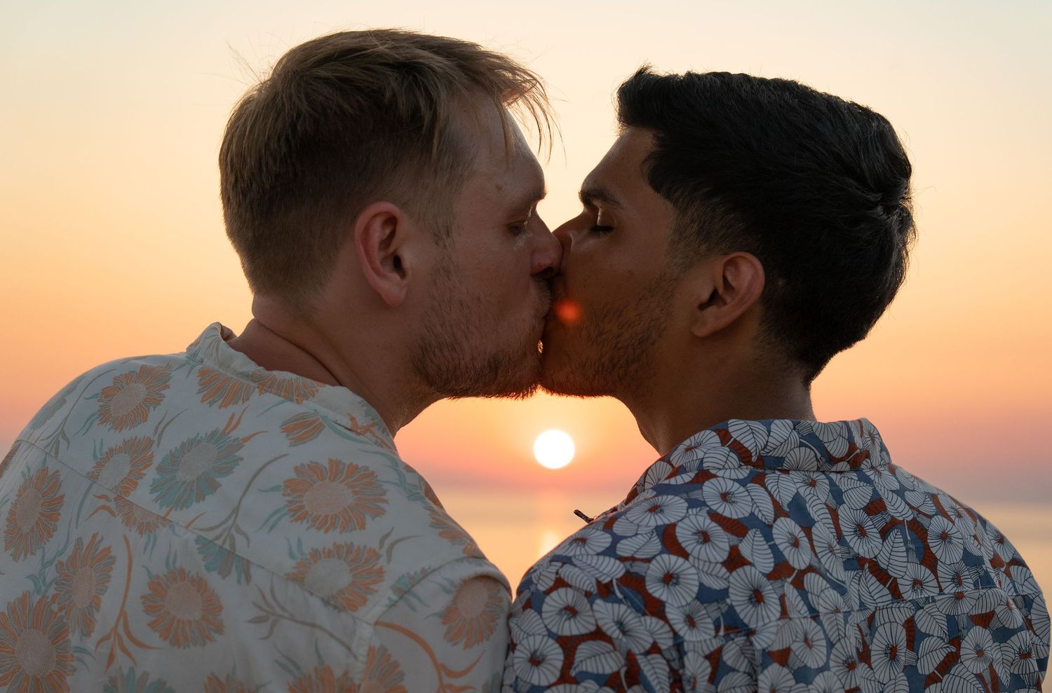 Two men sharing a romantic kiss against a stunning golden sunset over the ocean.