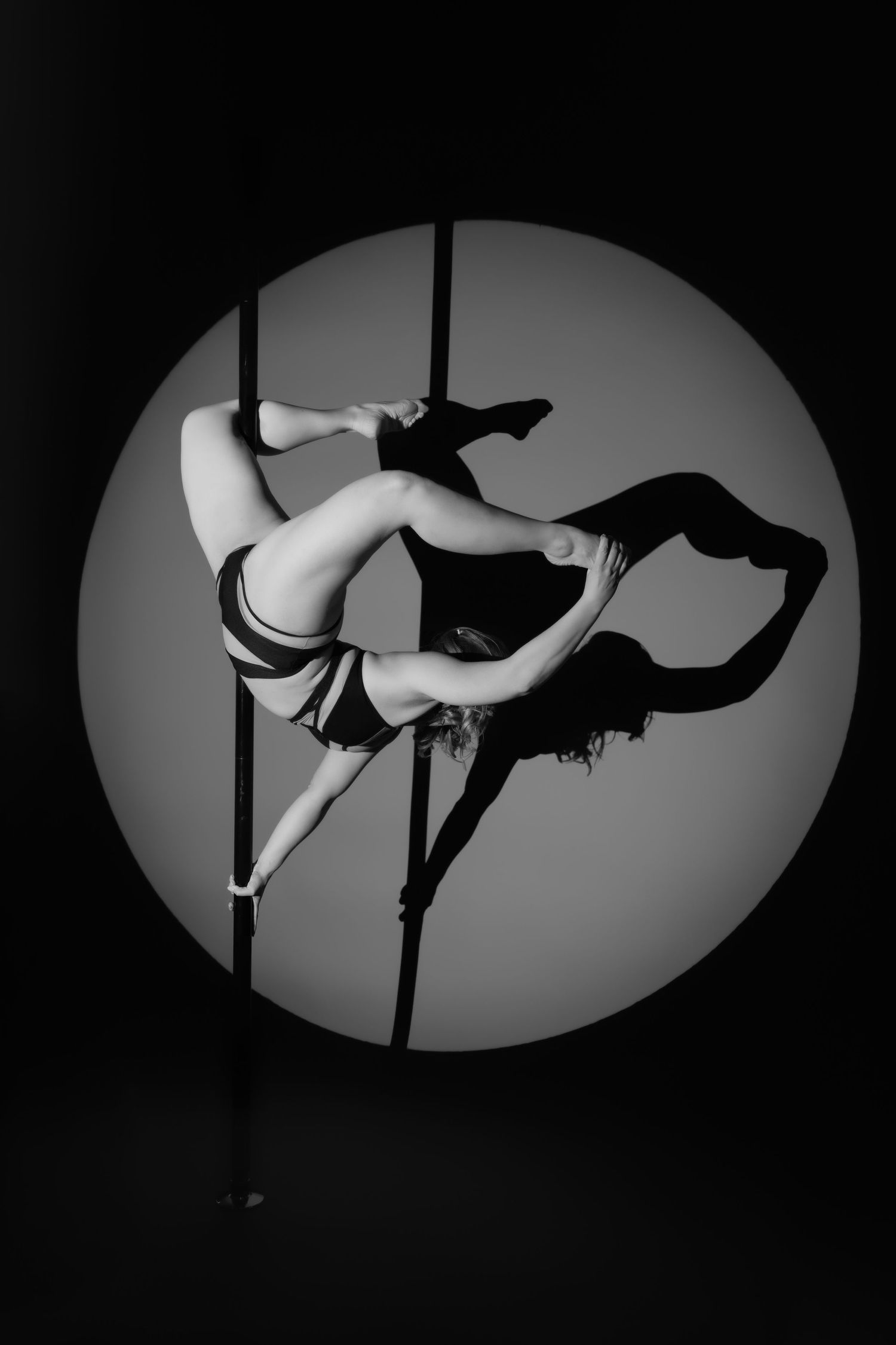 Black and white photo of a female pole dancer performing an inverted split pose in front of a circular spotlight.