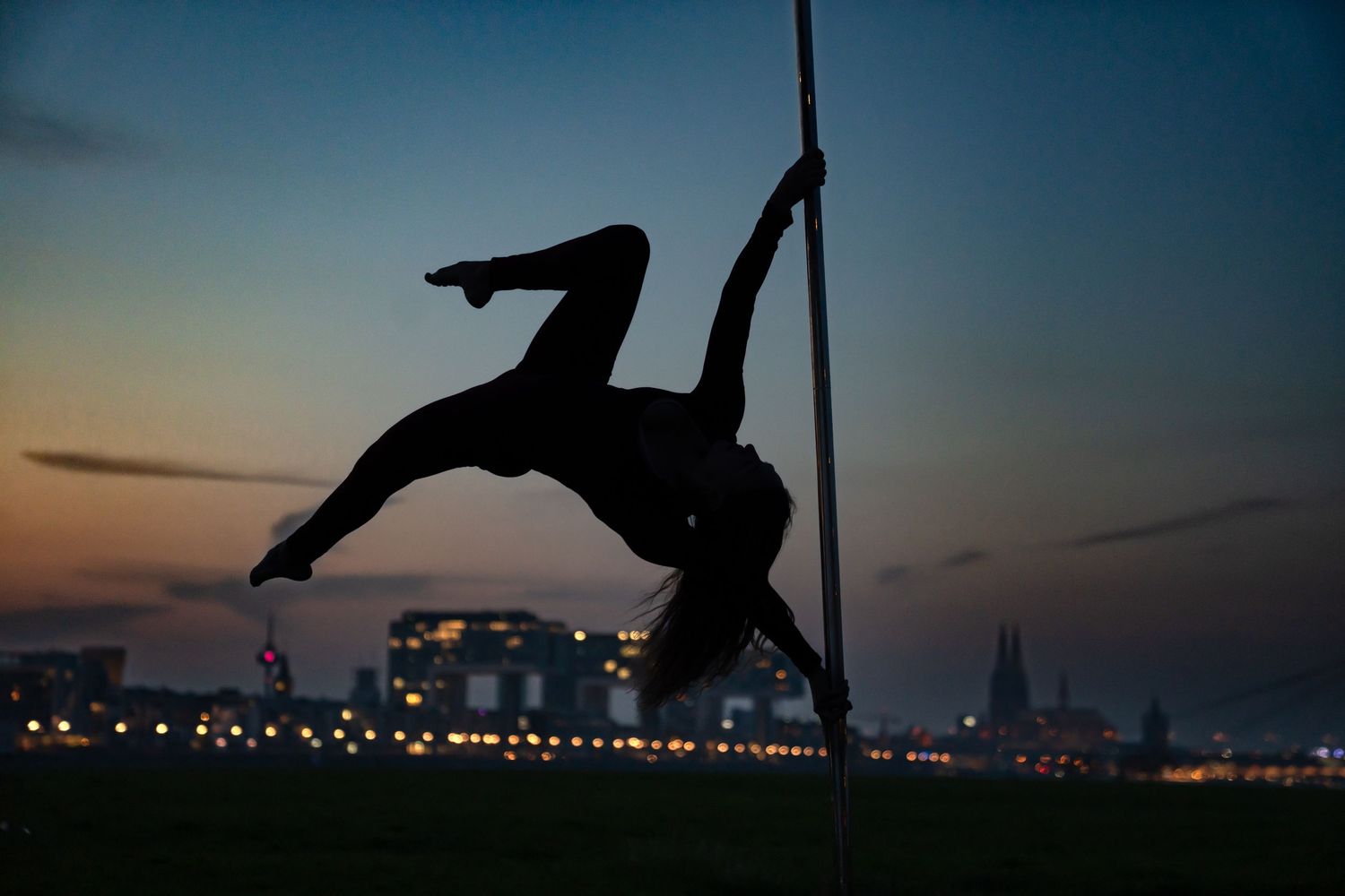 Silhouette of a pole dancer performing an athletic pose against a twilight city skyline backdrop.
