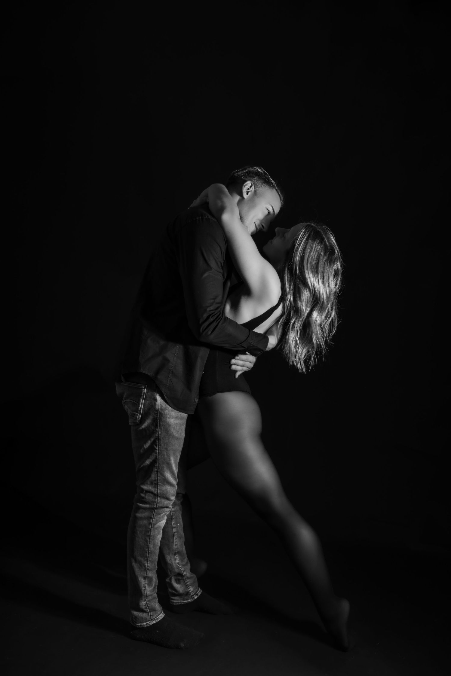 Black and white photo of a couple sharing a passionate kiss in a dramatic dark studio setting.