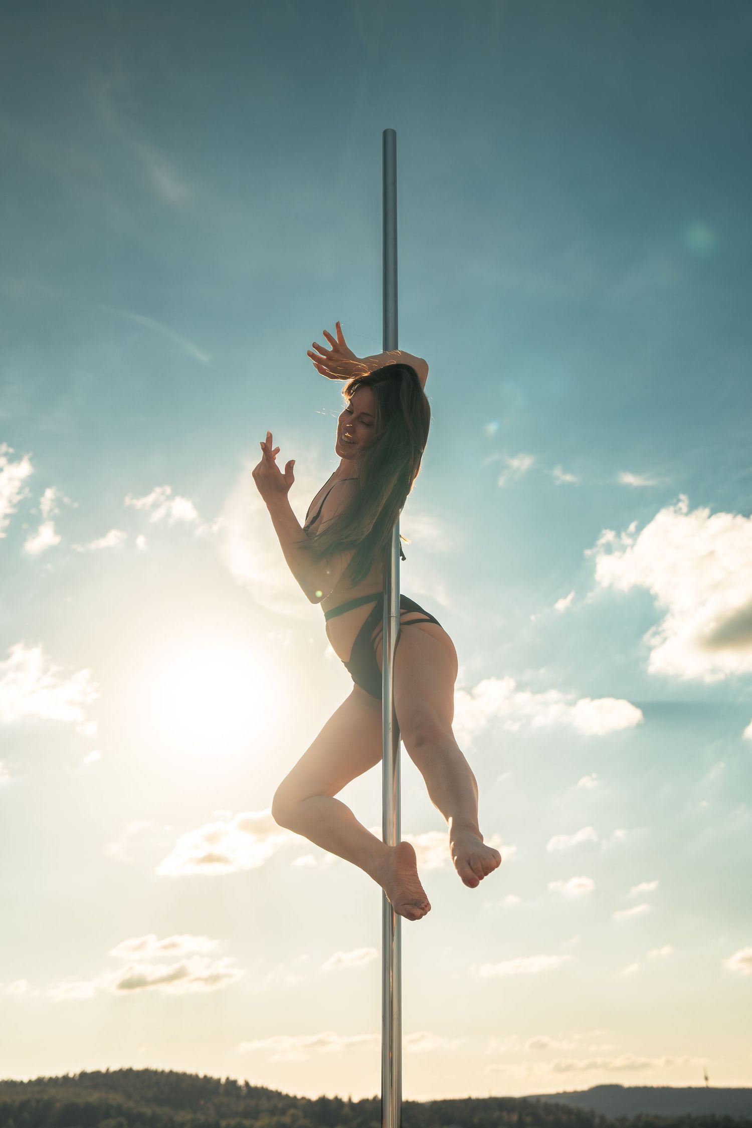 Woman performing pole dance outdoors against a blue sky with sunlight, showcasing athletic grace and flexibility.