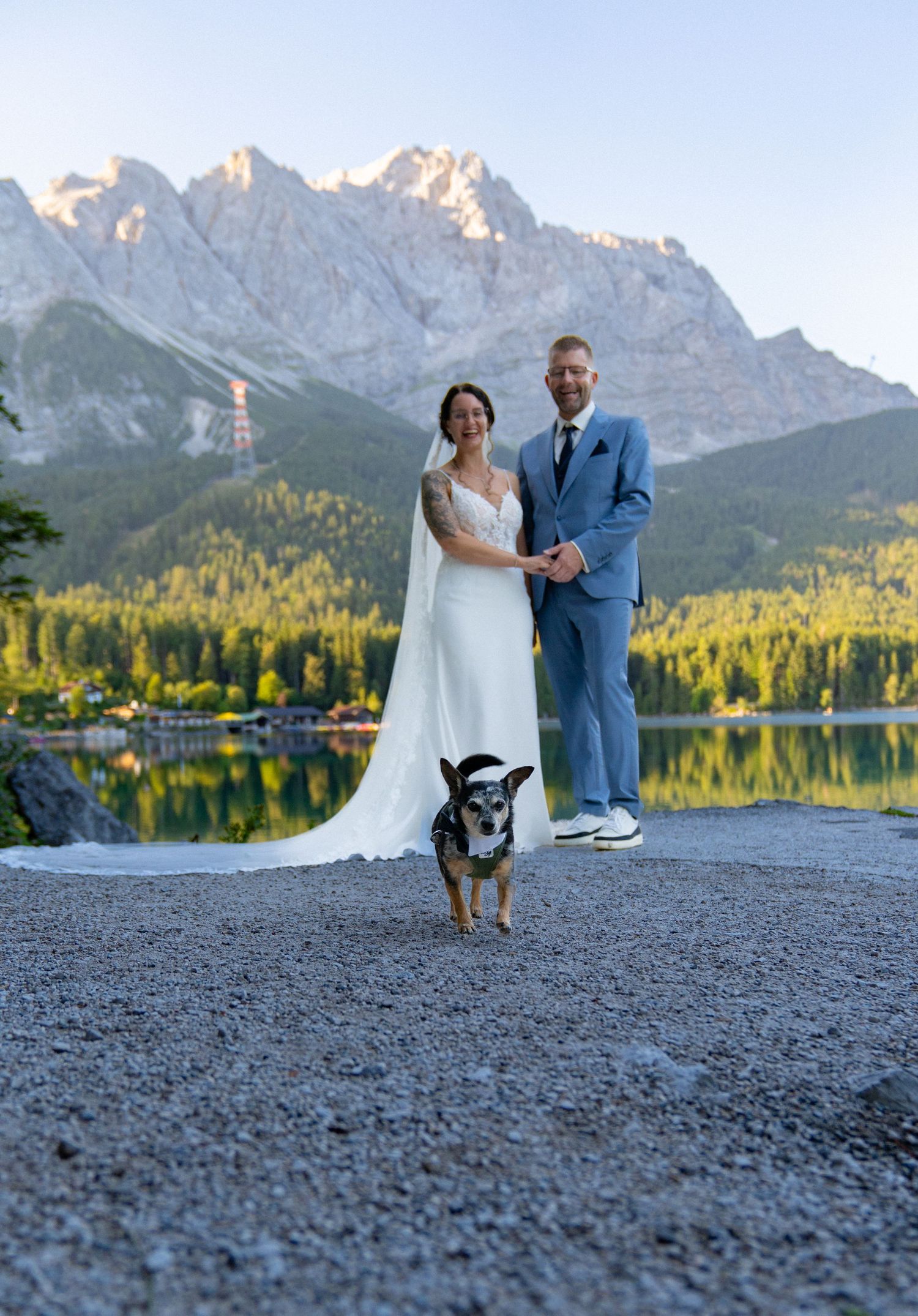 A couple in wedding attire poses with their dog against a scenic mountain lake backdrop at sunset.
