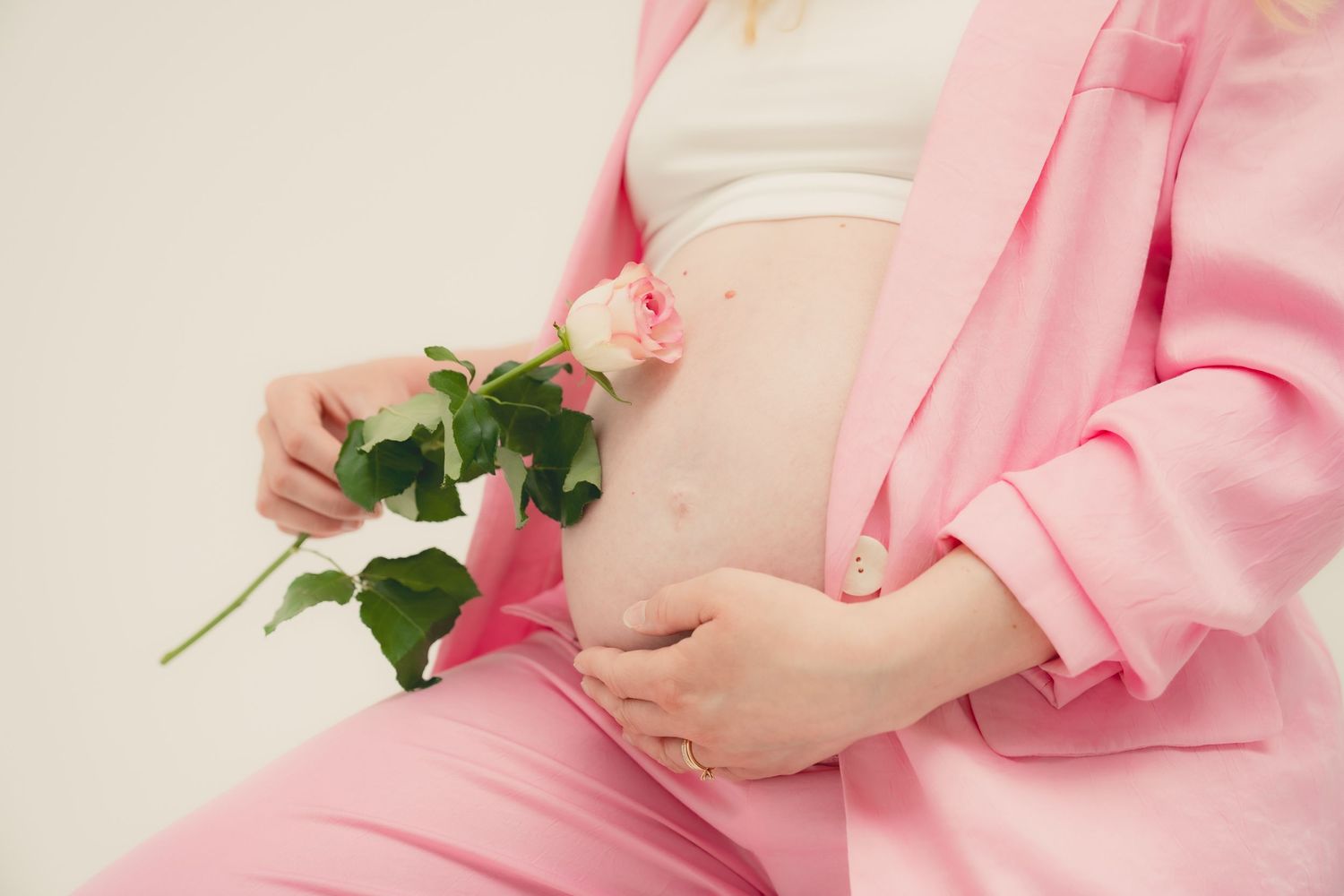 Pregnant woman in pink suit holding a pink rose against her bare baby bump on a light background.