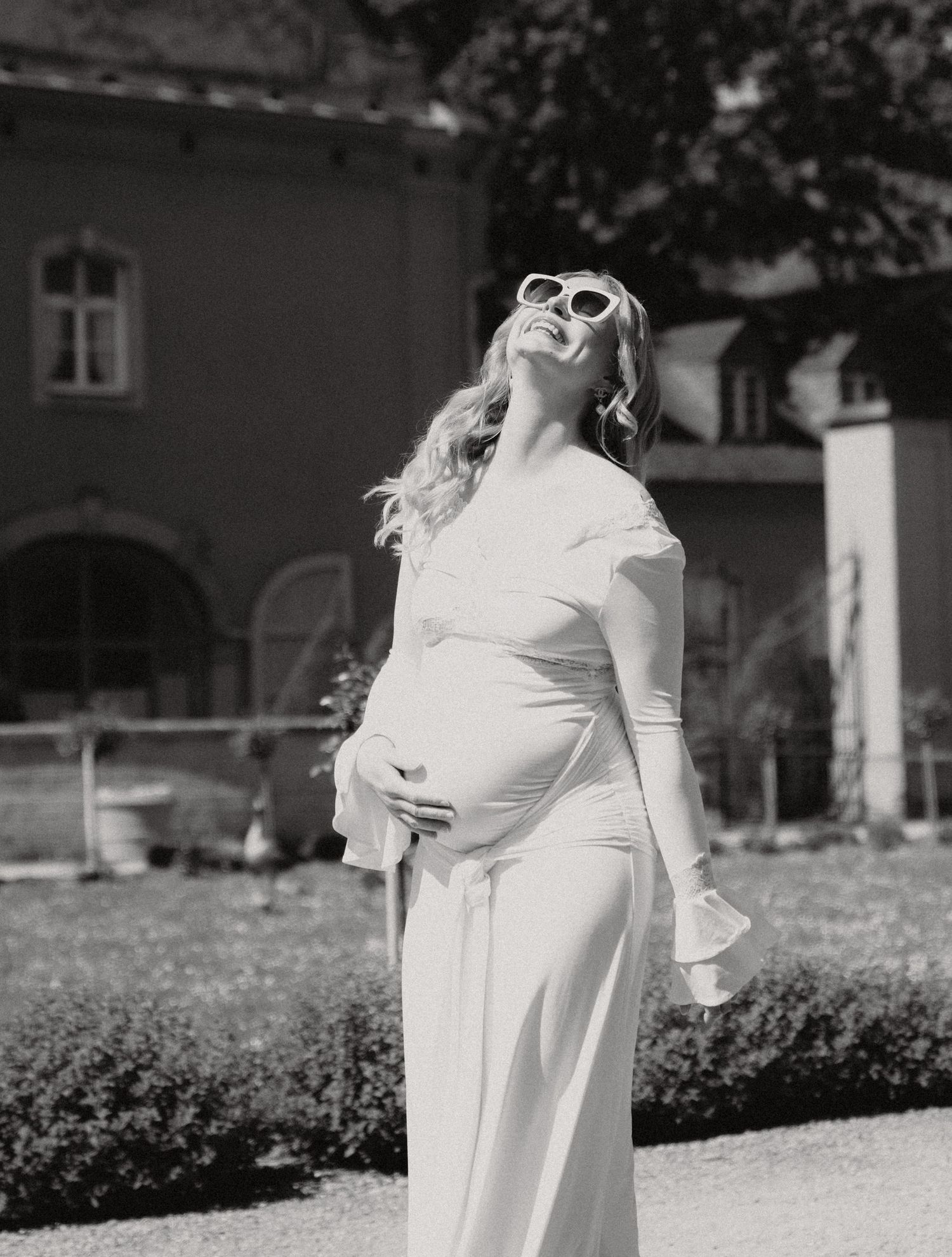 Black and white portrait of a pregnant woman in white dress and sunglasses posing outdoors.