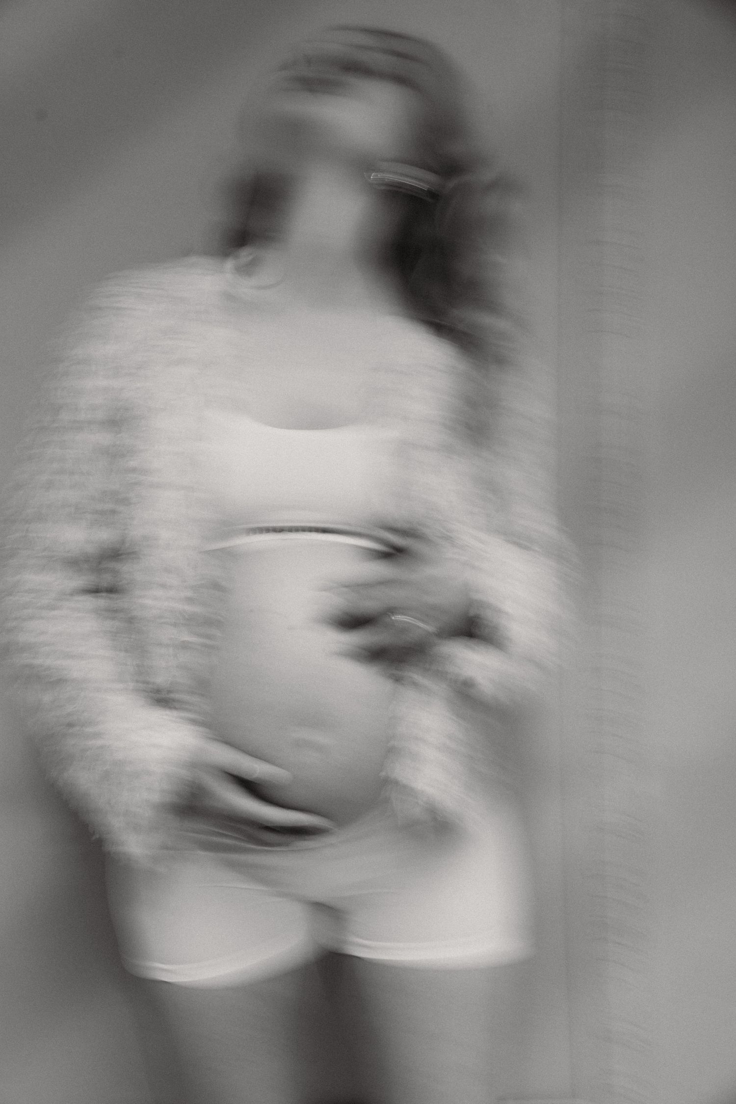 Black and white blurred photo of a pregnant woman cradling her baby bump, wearing white shorts and a fuzzy cardigan.