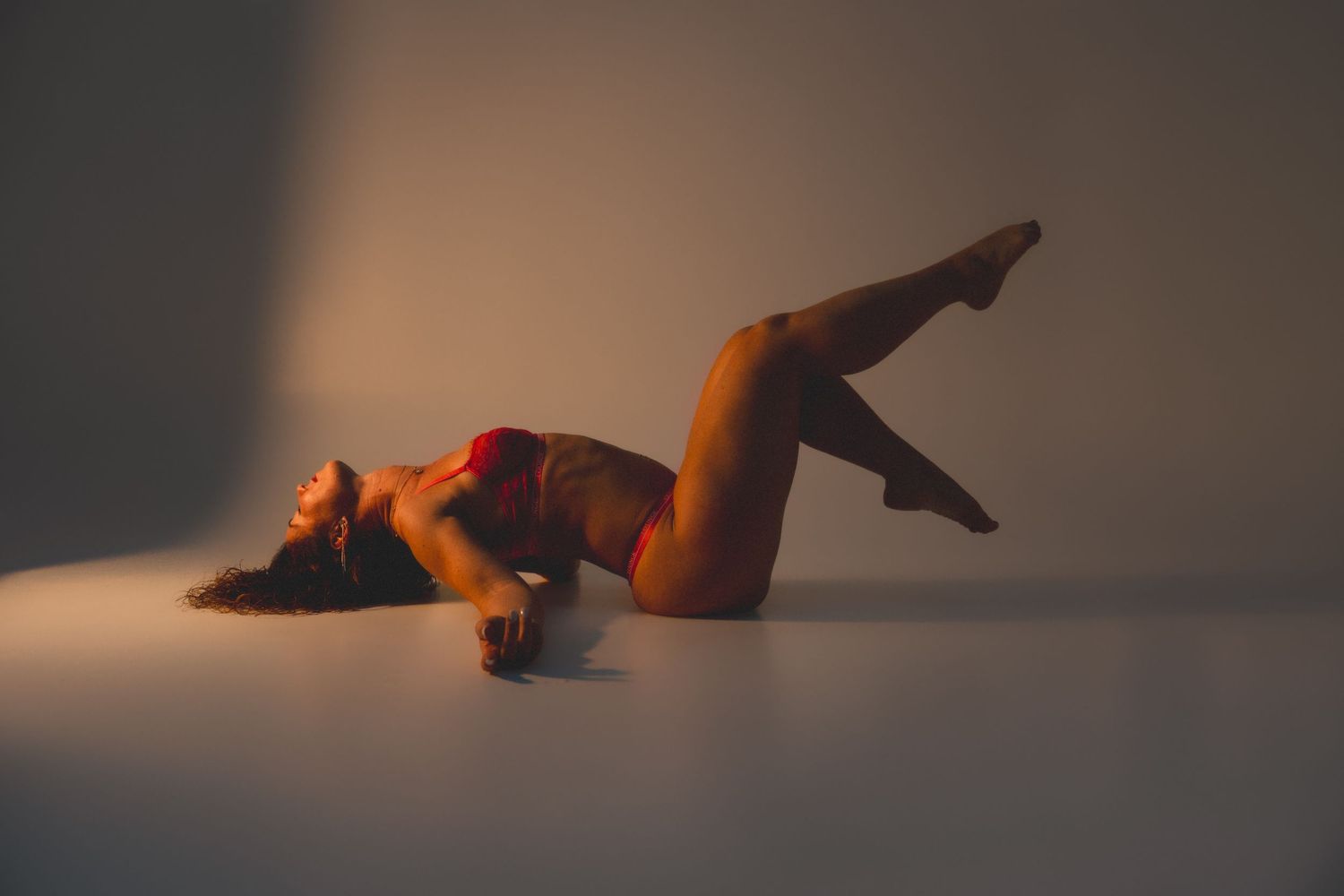 Woman in red lingerie posing dramatically on floor in warm golden light against neutral background.