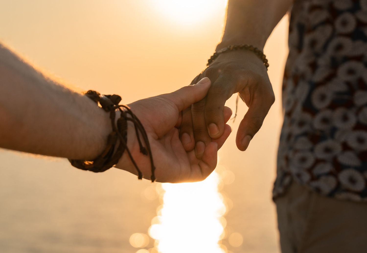 Couple holding hands at sunset near water, wearing matching bracelets, romantic golden hour moment.