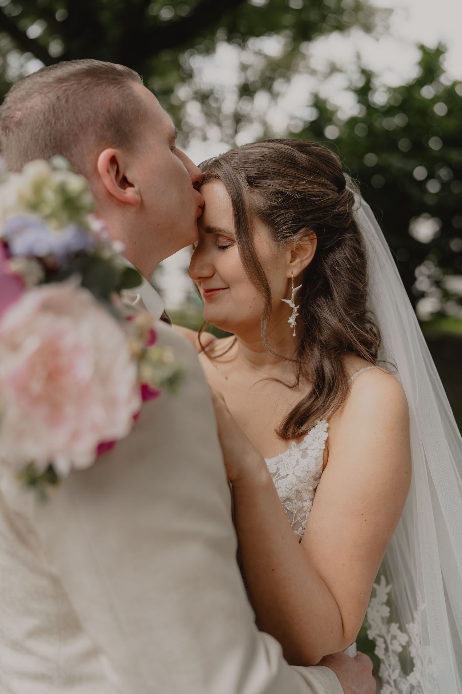 Groom kissing bride on forehead during outdoor wedding, bride smiling softly in lace dress and veil holding pink bouquet.