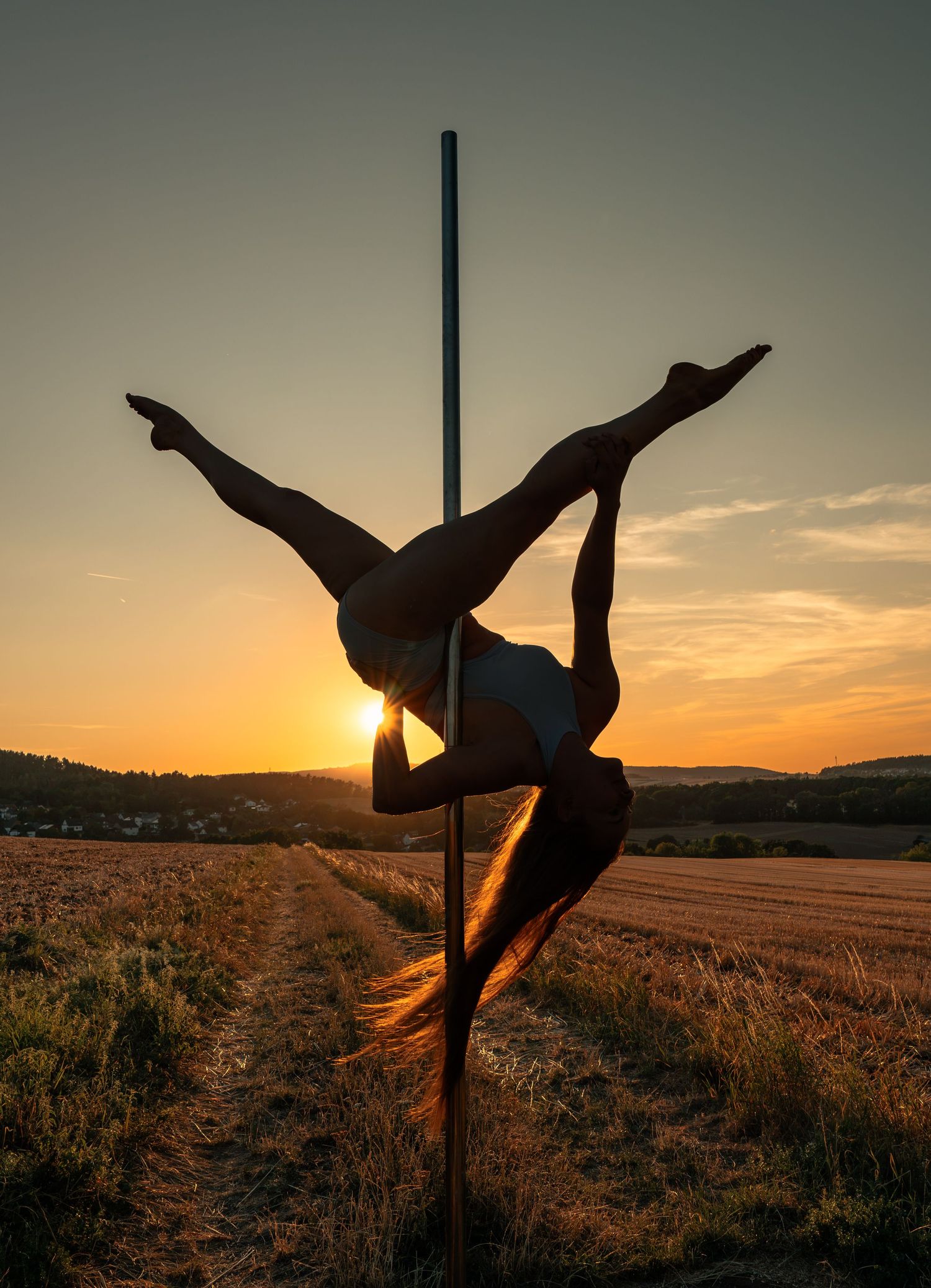 Silhouette of a pole dancer performing an acrobatic split outdoors at golden sunset over a rural field.
