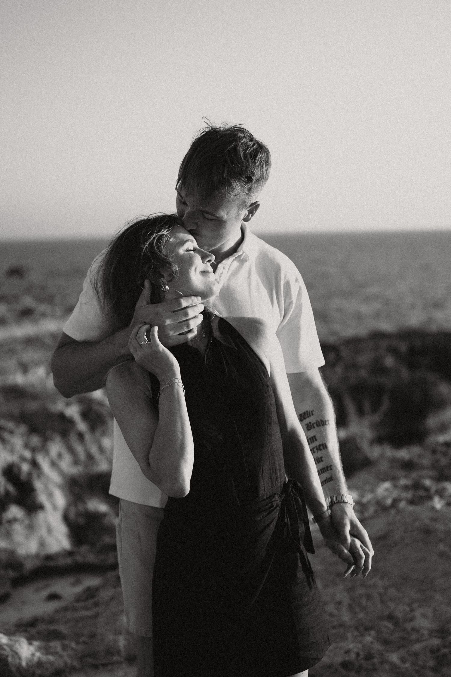 Black and white photo of a couple embracing on rocky coastal cliffs, man kissing womans cheek at sunset.