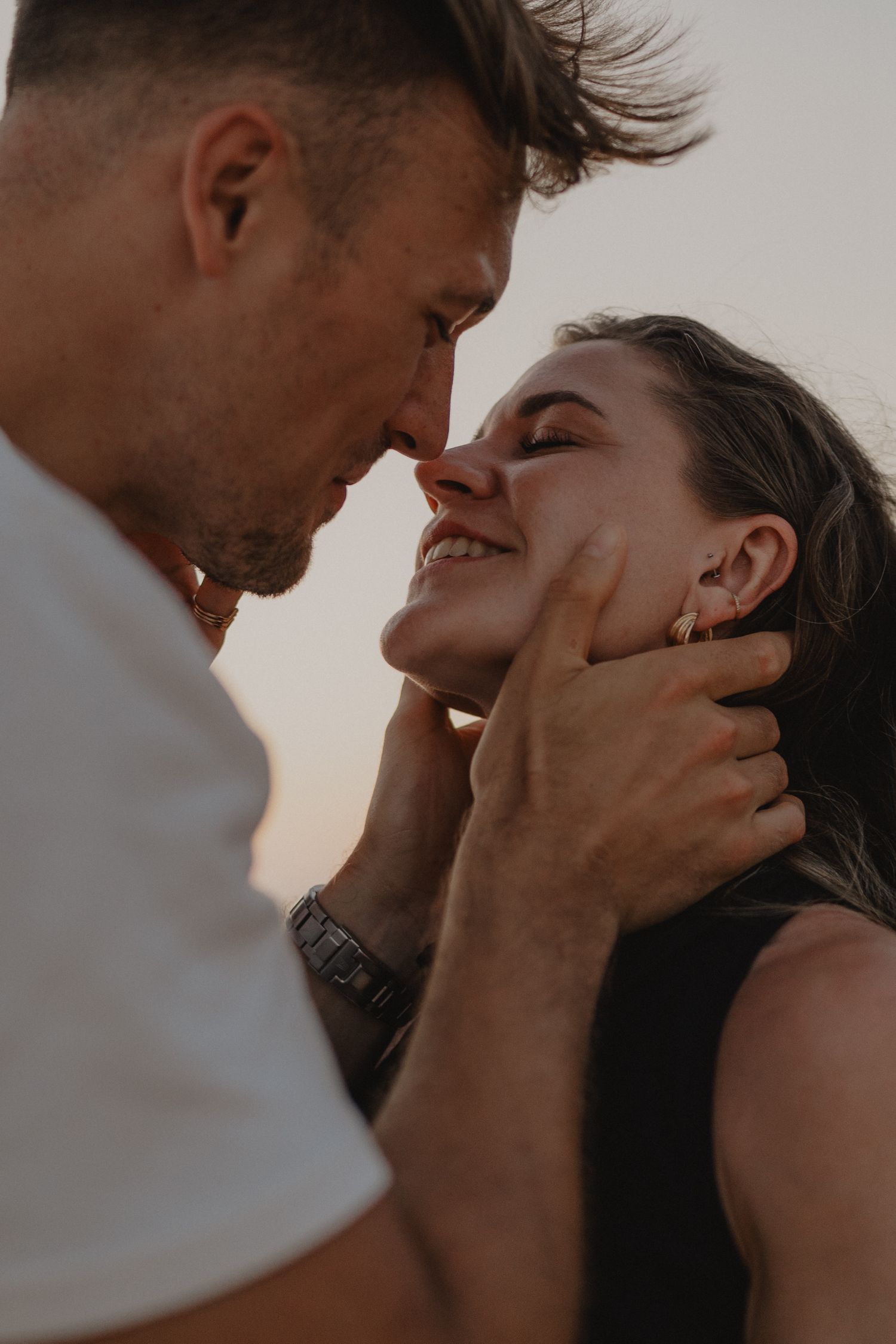 A couple shares a romantic moment, the man gently cupping the smiling woman's face as they lean in for a kiss at sunset.