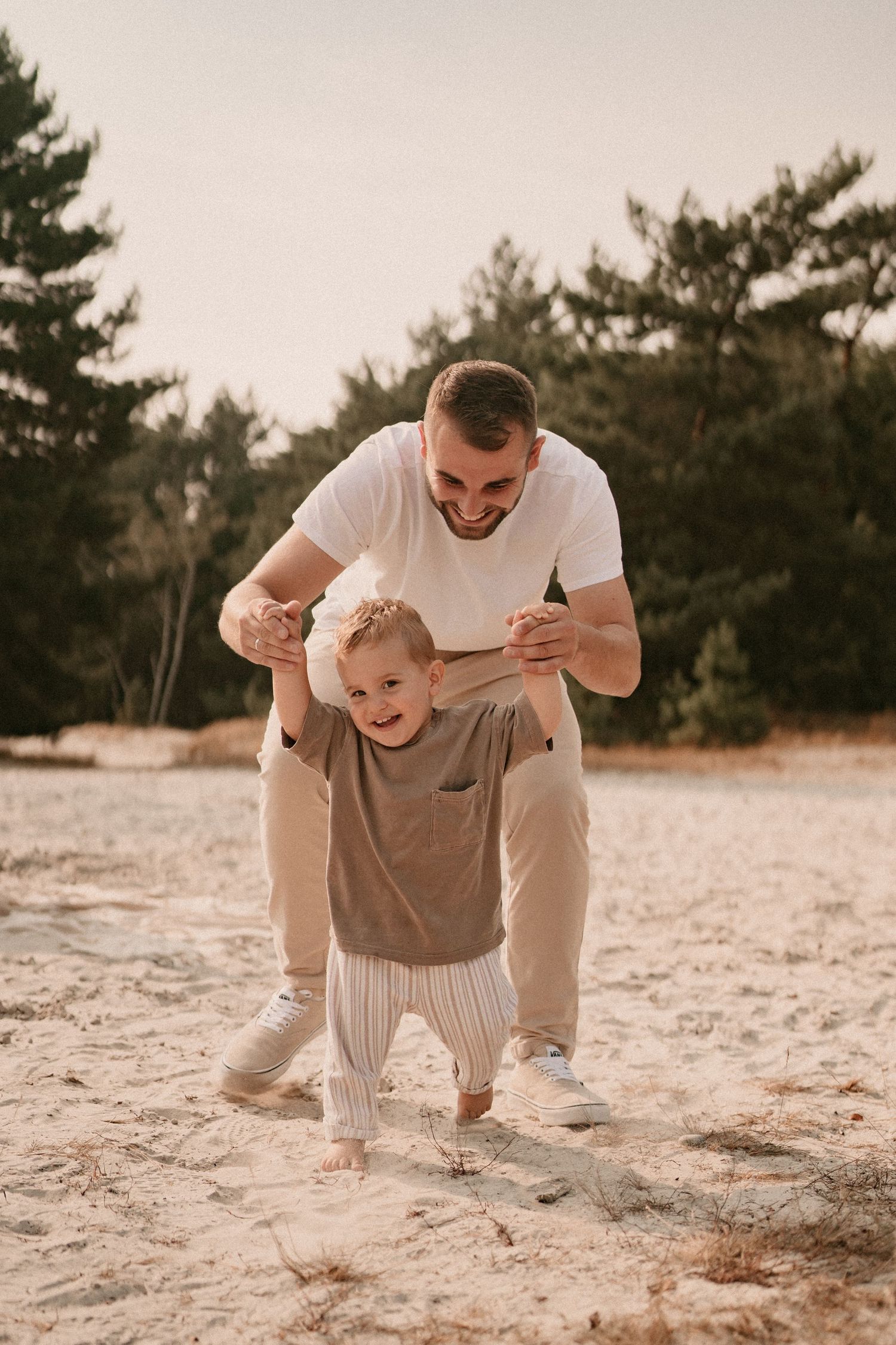 A playful sequence of a parent and toddler crawling together on sandy ground with pine trees in the background.