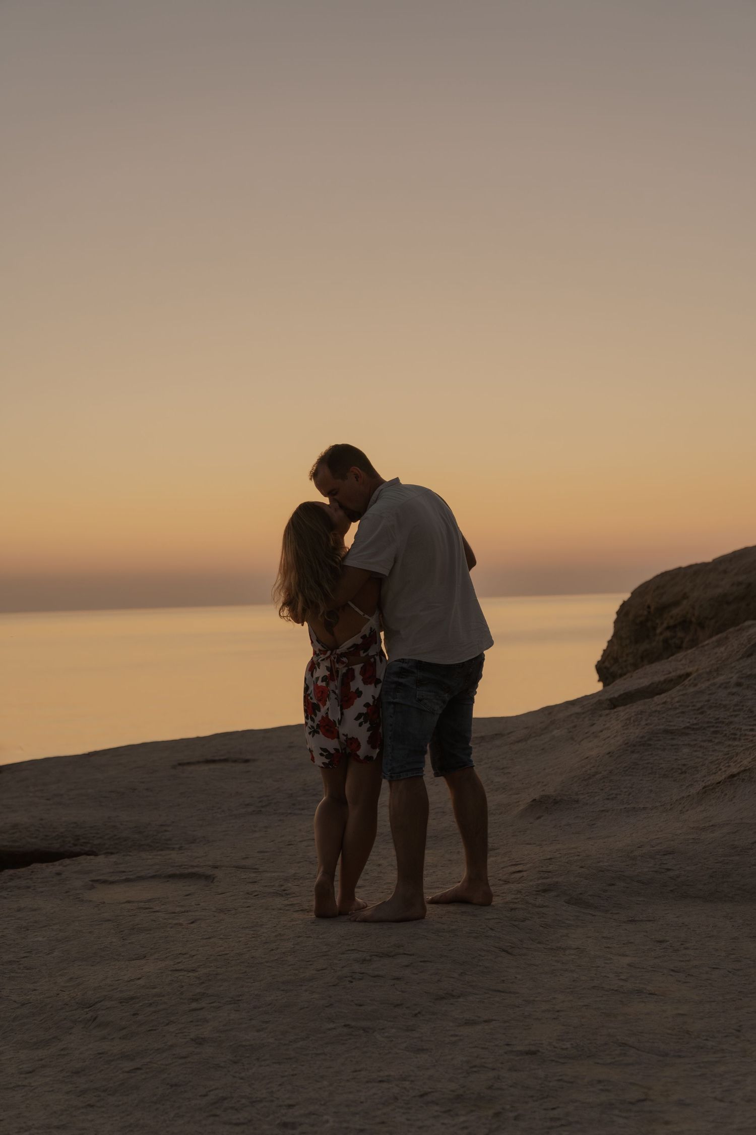 Silhouetted couple sharing intimate moment on rocky beach during sunset.