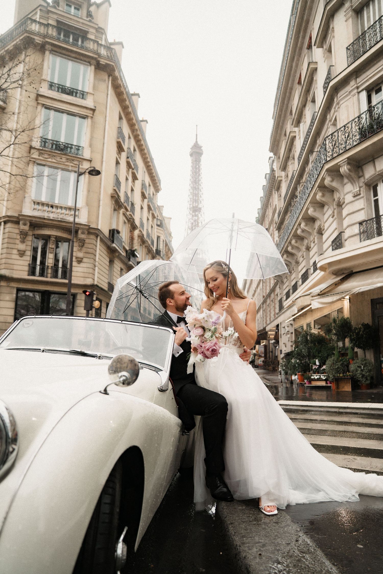 Wedding couple poses with vintage white car on cobblestone streets of Paris with Eiffel Tower in background.