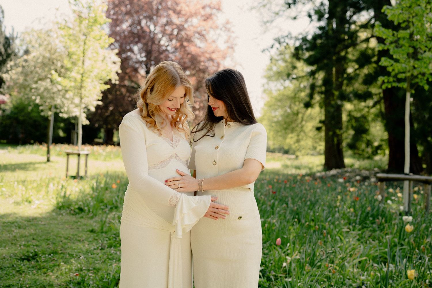 Two pregnant women in cream dresses tenderly cradling their baby bumps together in a sunny garden.