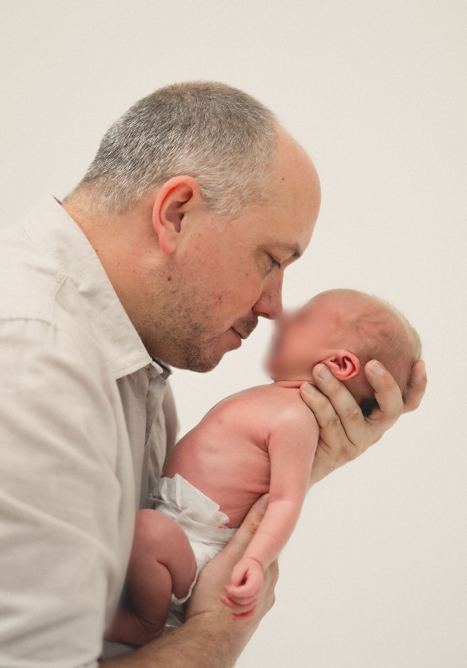 A father tenderly kisses his newborn baby, cradling the infant gently in both hands against a white background.