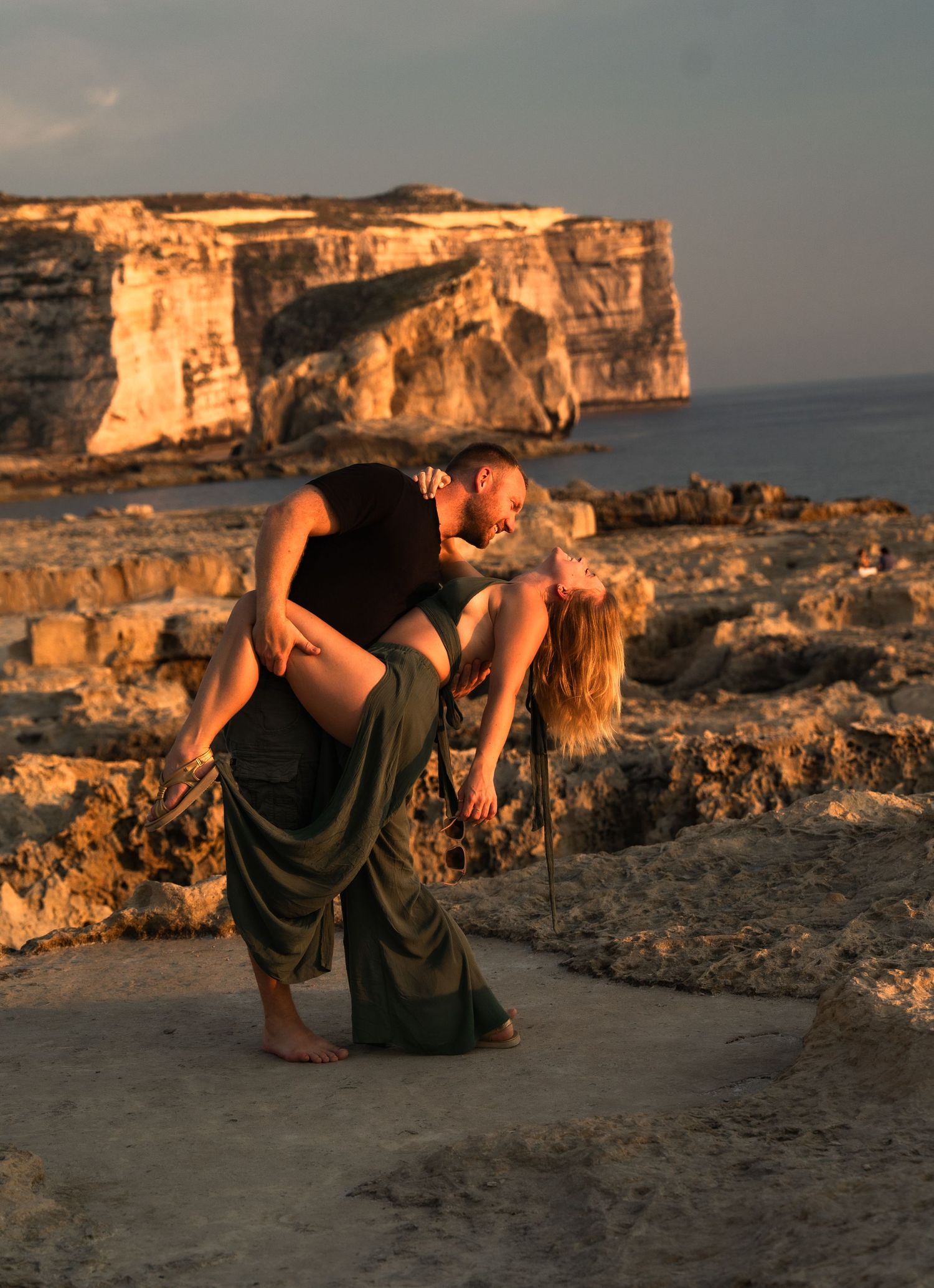 Couple sharing a romantic dip kiss on rocky coastal cliffs at golden sunset with dramatic sea cliffs in background.