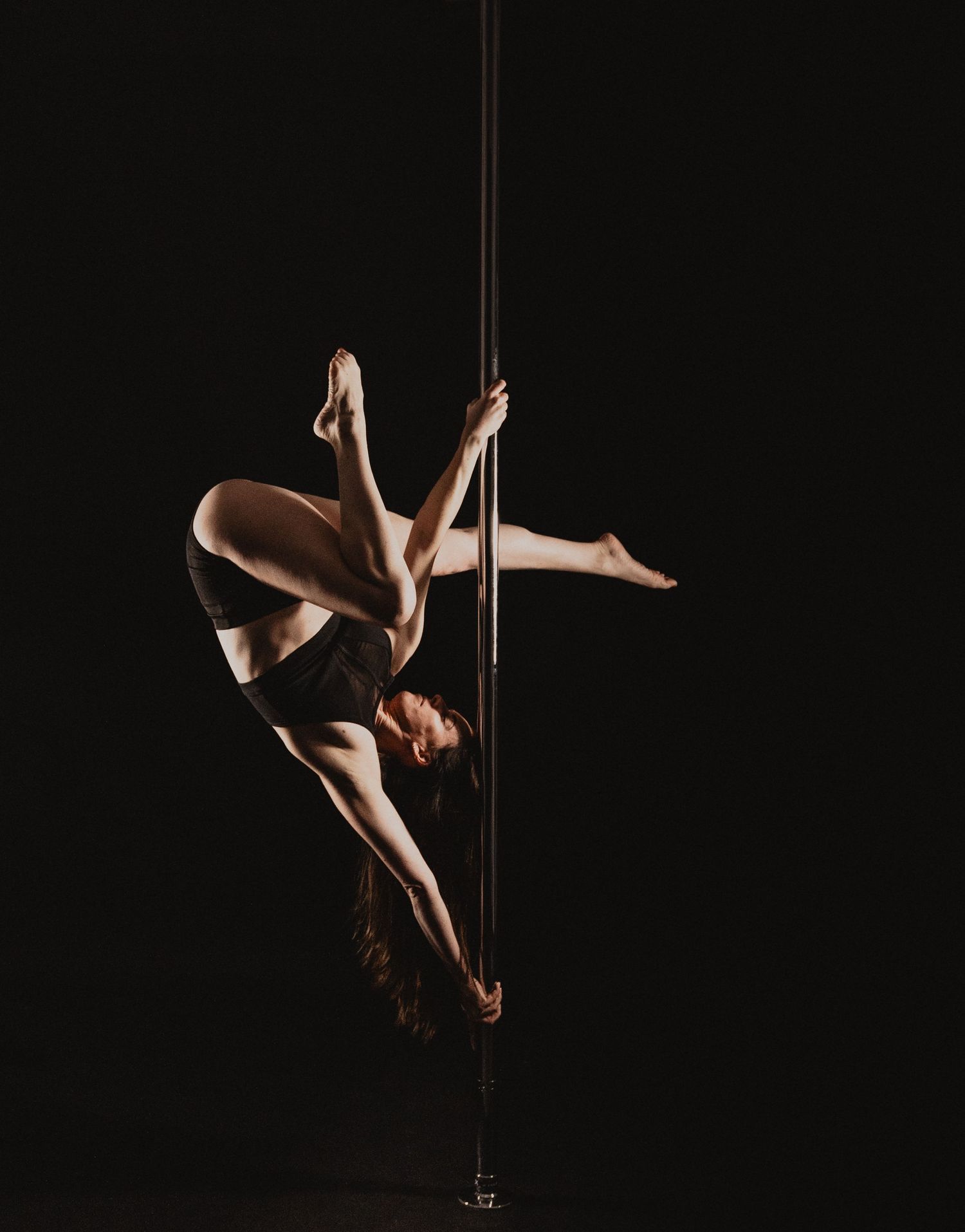 Flexible pole dancer performs an inverted split on a vertical pole against a dark black background.
