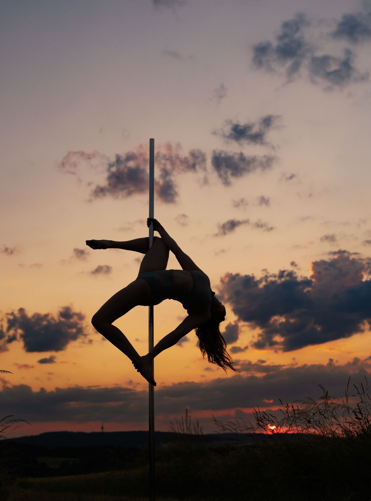 Silhouette of a woman performing pole dance against a dramatic sunset sky with glowing orange and purple clouds.