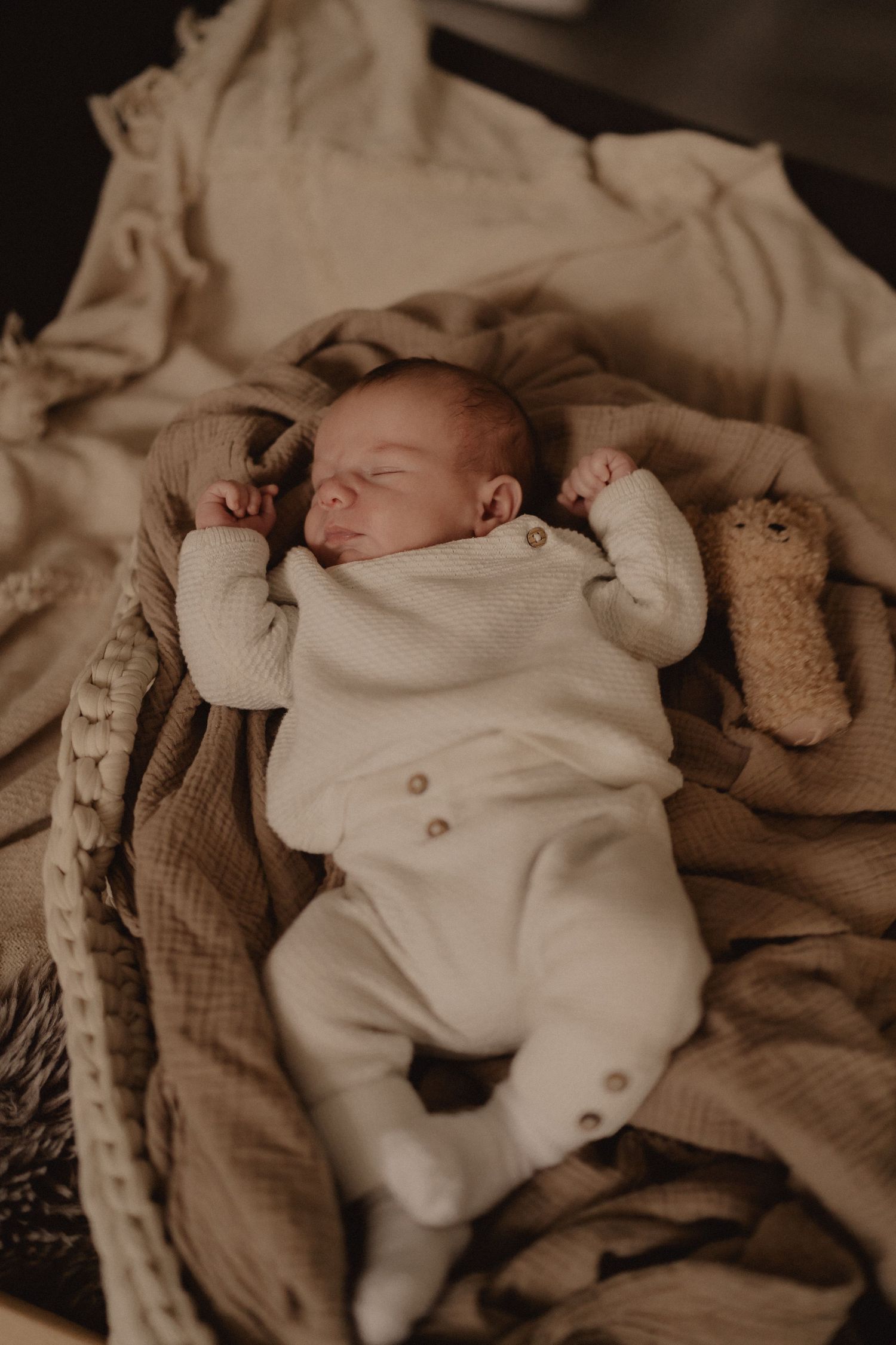 Baby in white onesie sleeping peacefully with teddy bear on beige blanket.