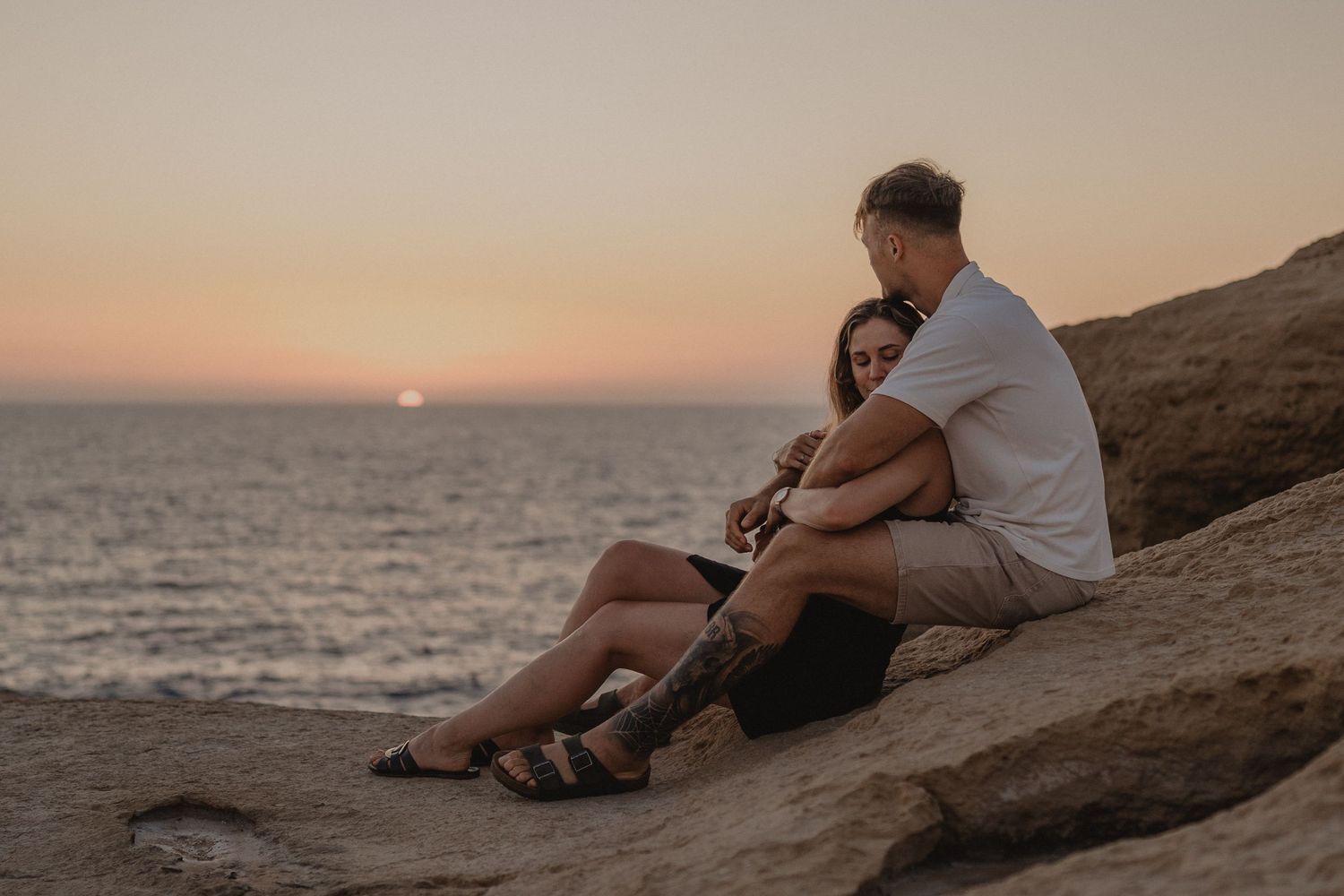 Couple sitting on rocky cliffs embracing while watching a romantic sunset over the ocean.