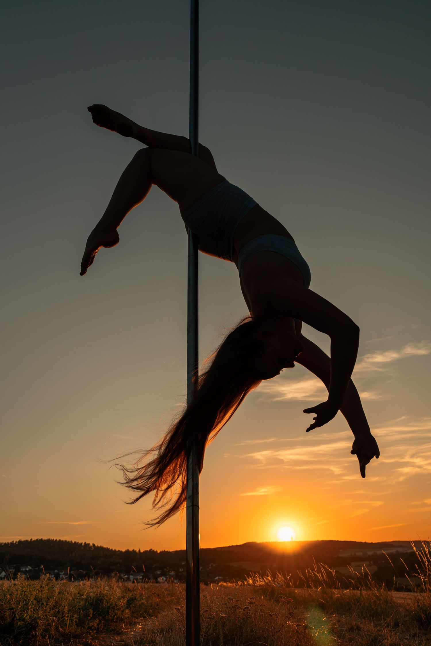Silhouette of a woman performing pole dance against a vibrant golden sunset sky outdoors.